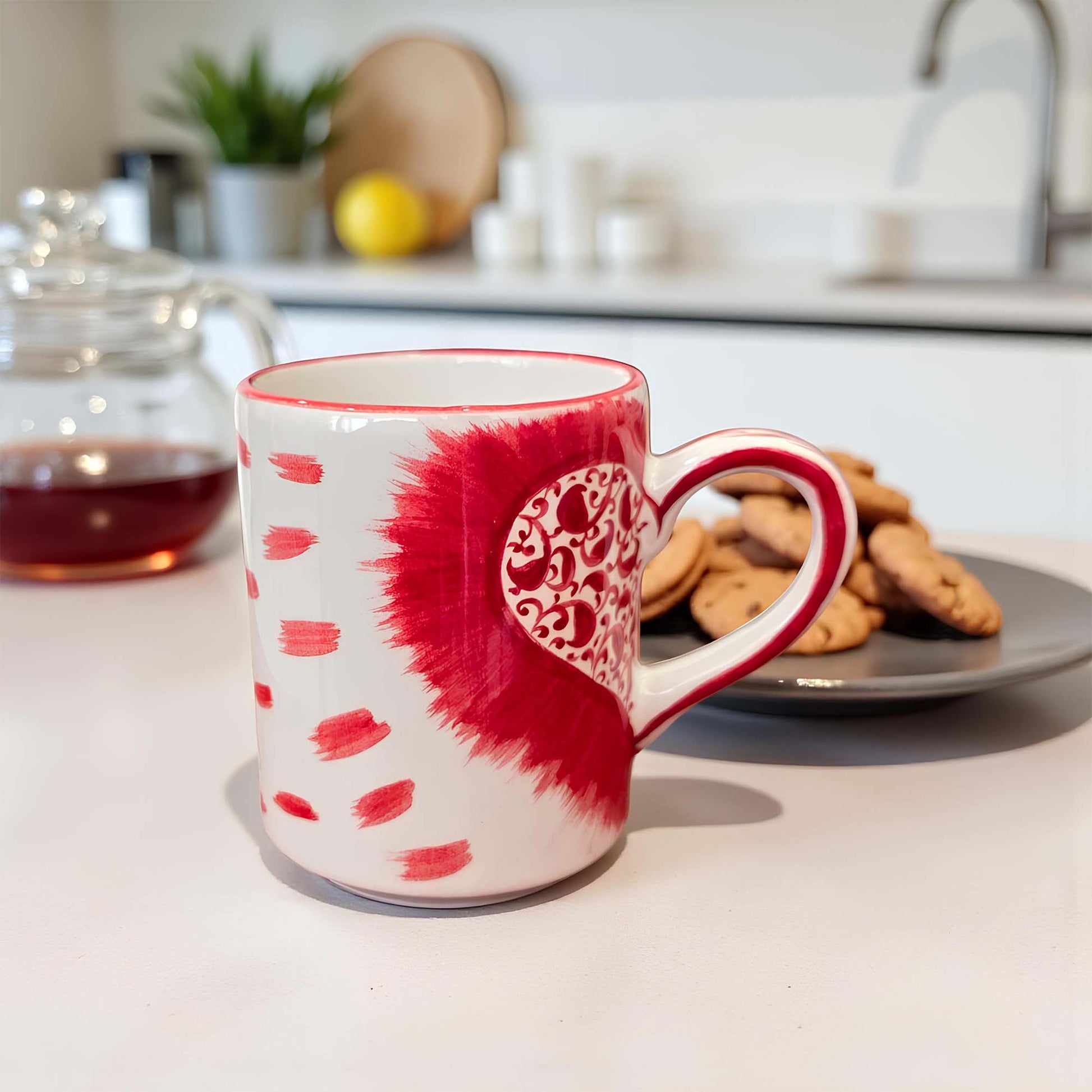 Red and white mug with heart design on a kitchen counter with cookies and tea.