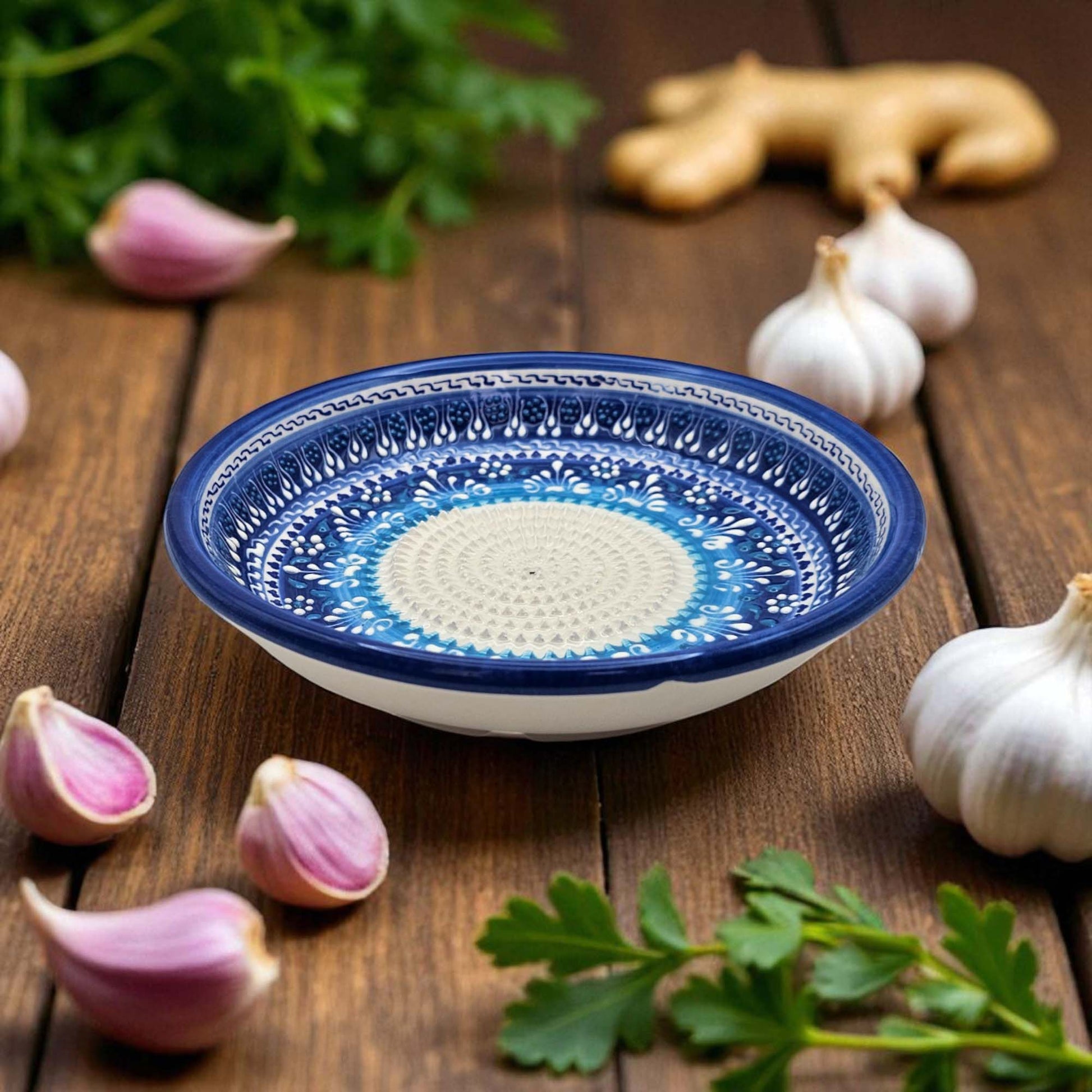 Blue and white decorative garlic grater bowl on a wooden surface with garlic and herbs.