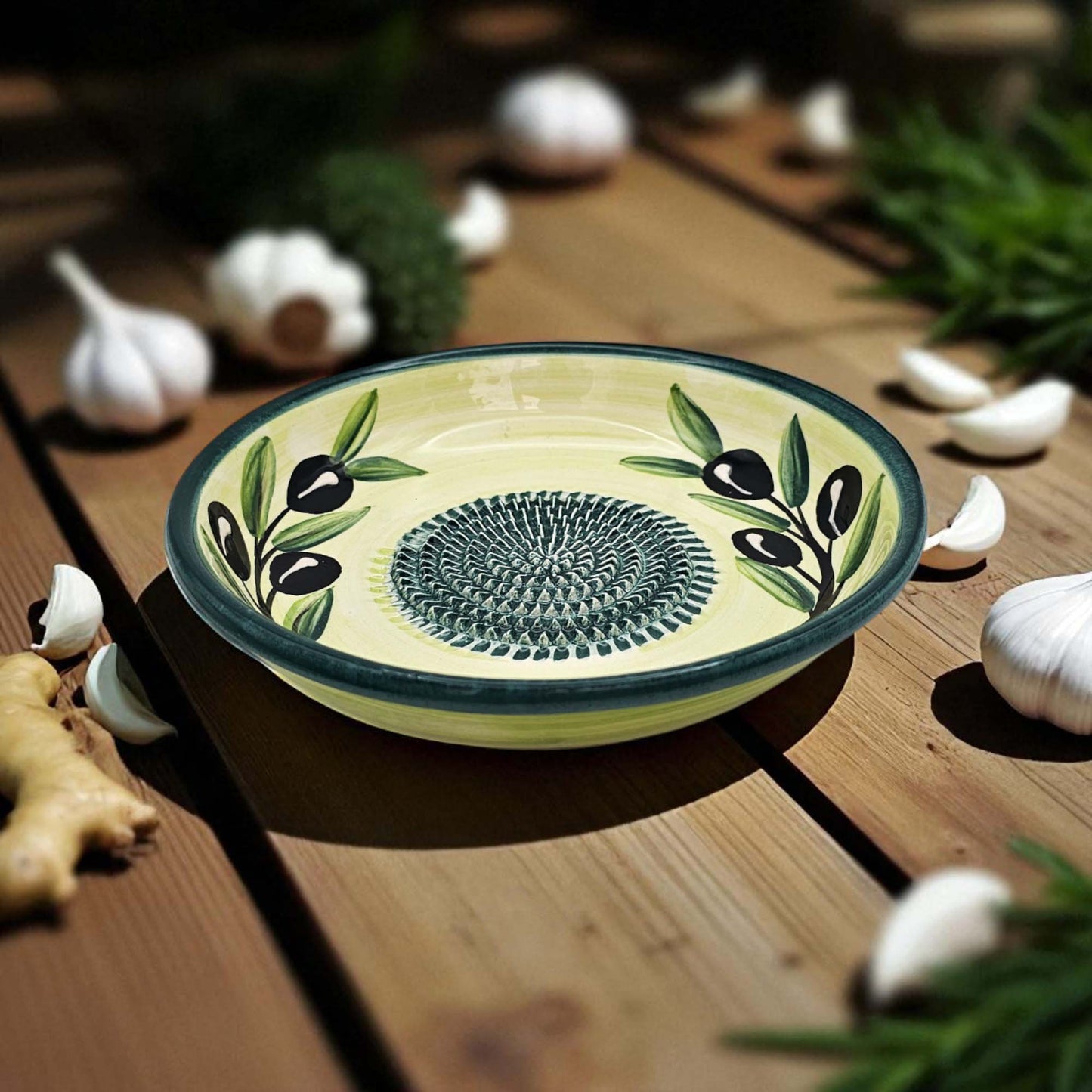 Ceramic garlic grater bowl with olive design on a wooden surface with garlic and herbs.