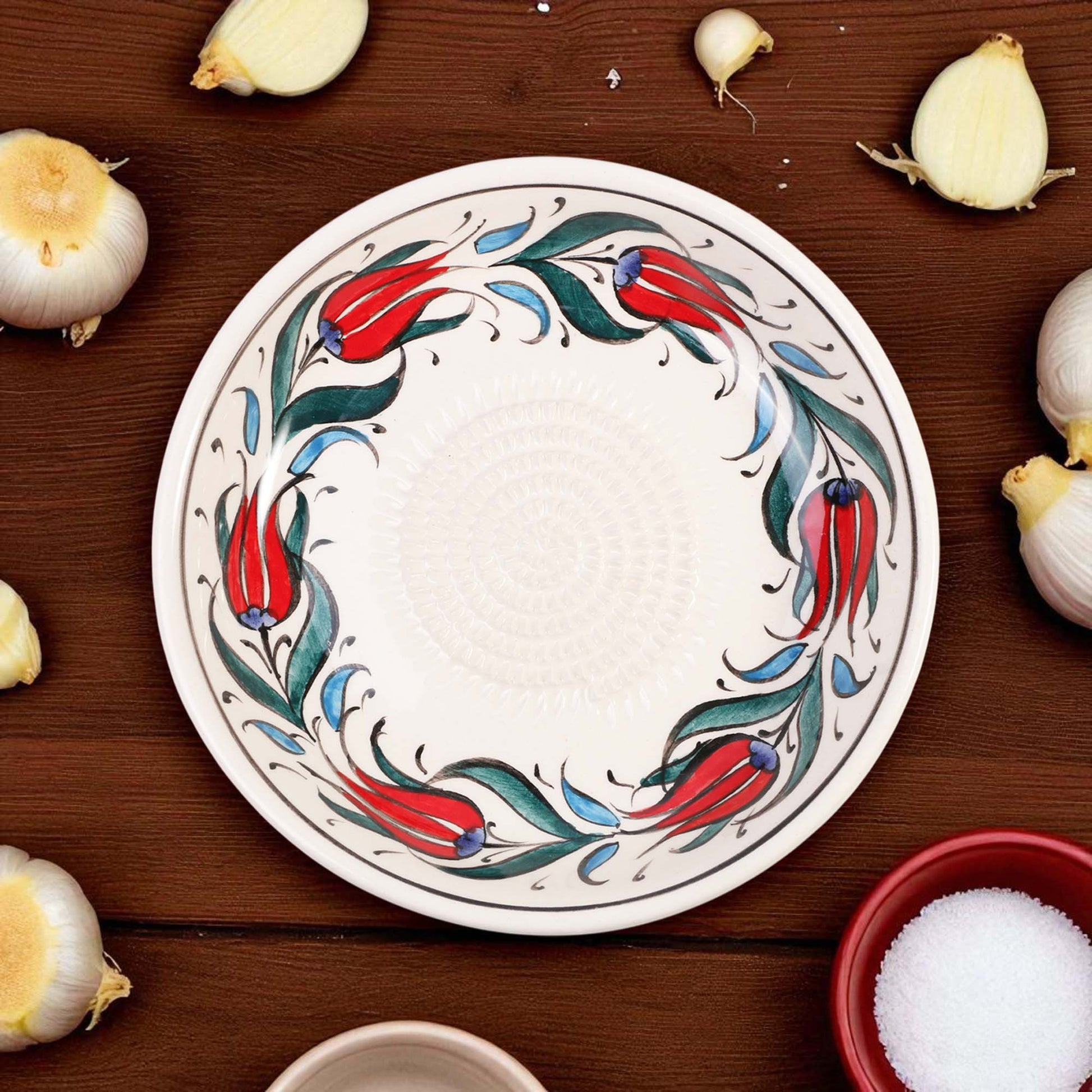 Decorative ceramic garlic grater bowl with floral design on a wooden surface with garlic bulbs.