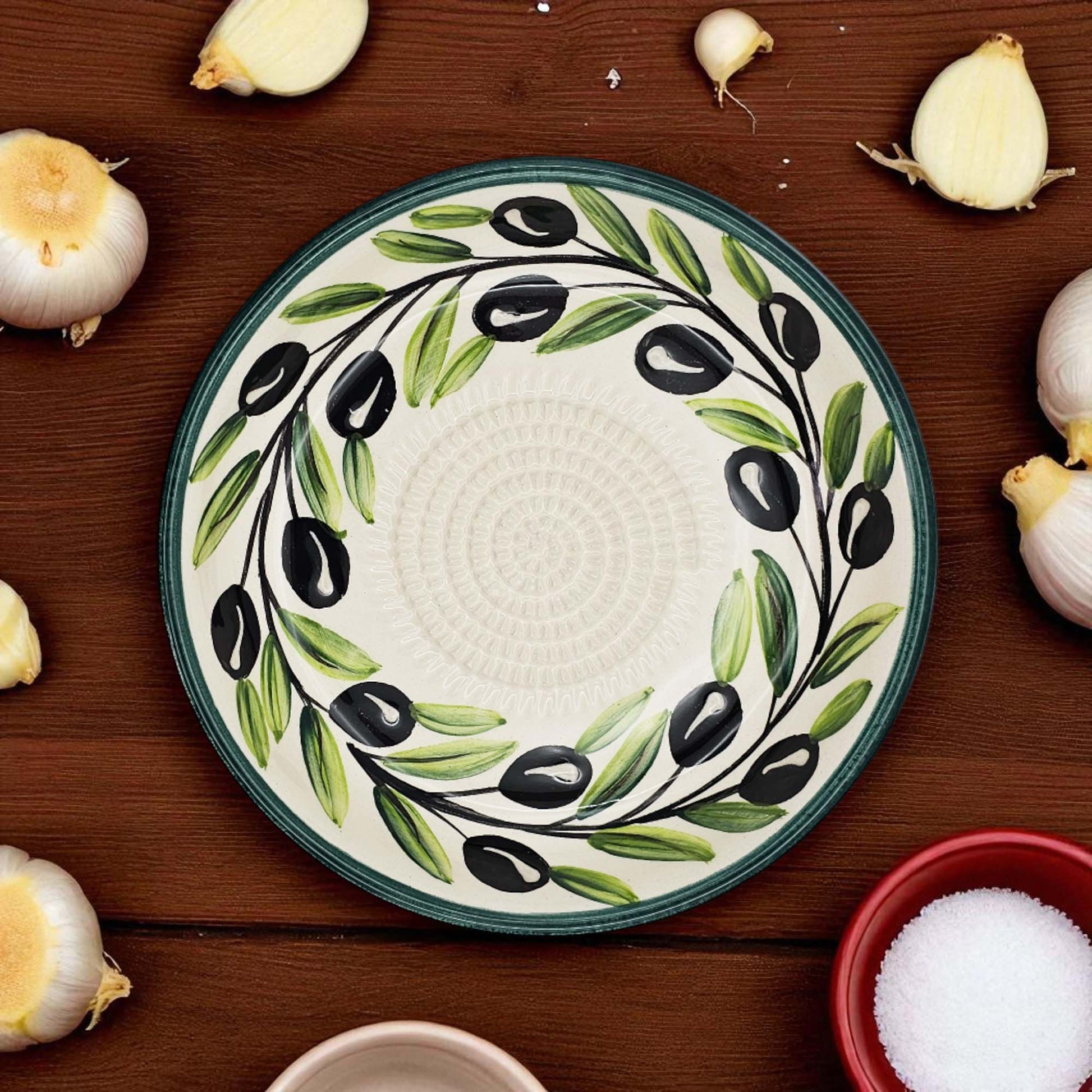 Decorative garlic grater bowl with olive design on a wooden surface with garlic bulbs and a bowl of salt.