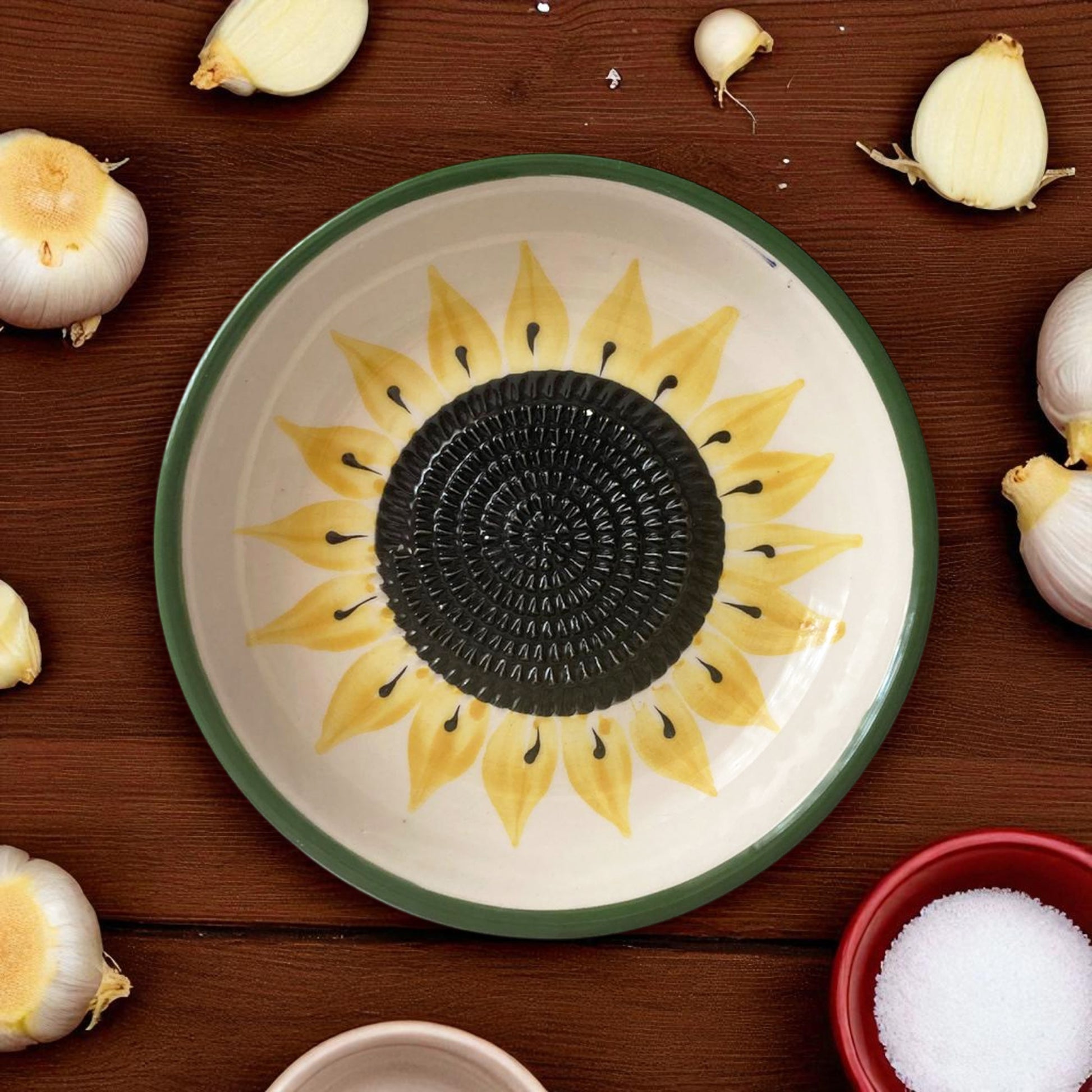 Ceramic garlic grater bowl with sunflower design on a wooden surface with garlic bulbs.