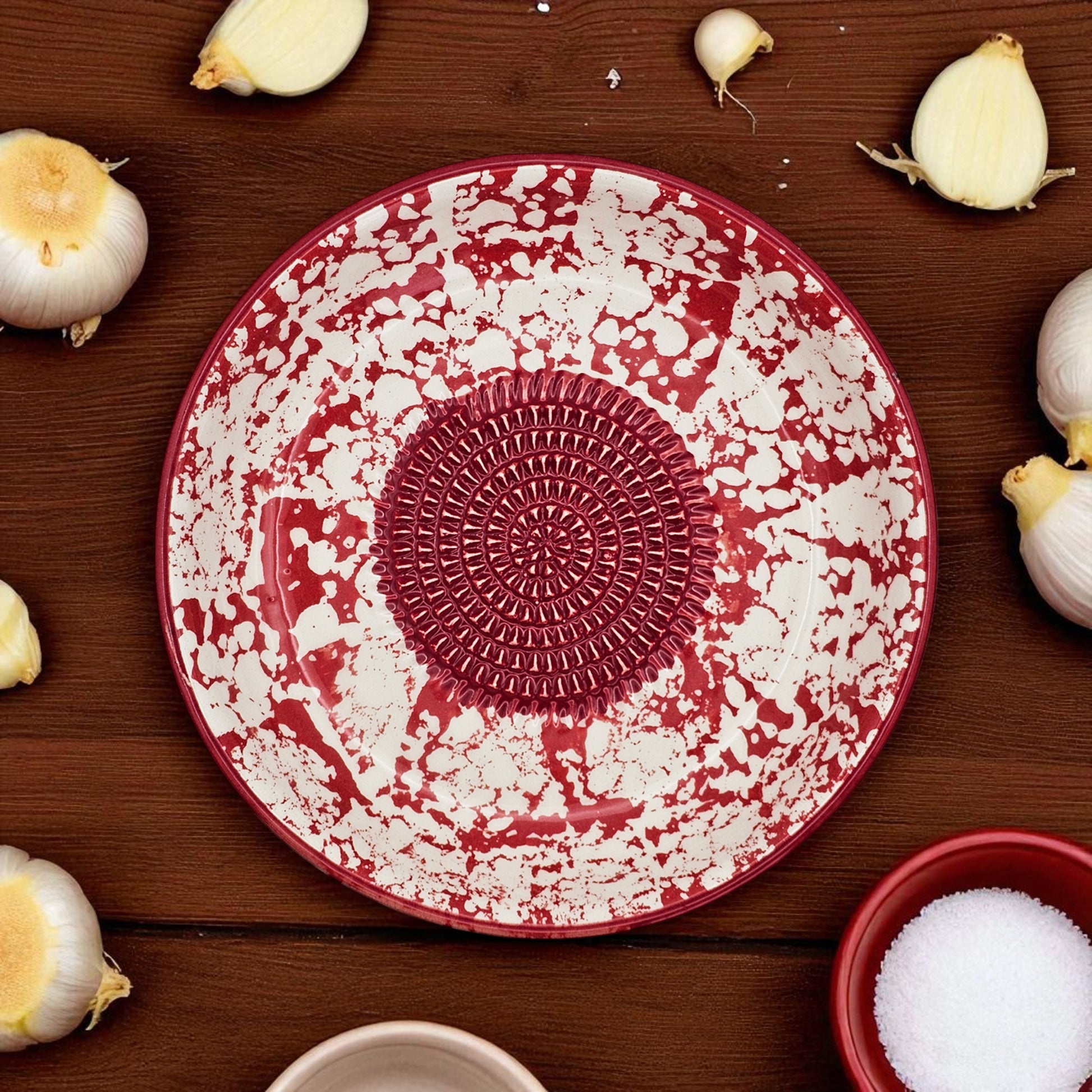 Red and white ceramic garlic grater bowl with decorative center on a wooden surface with garlic bulbs.
