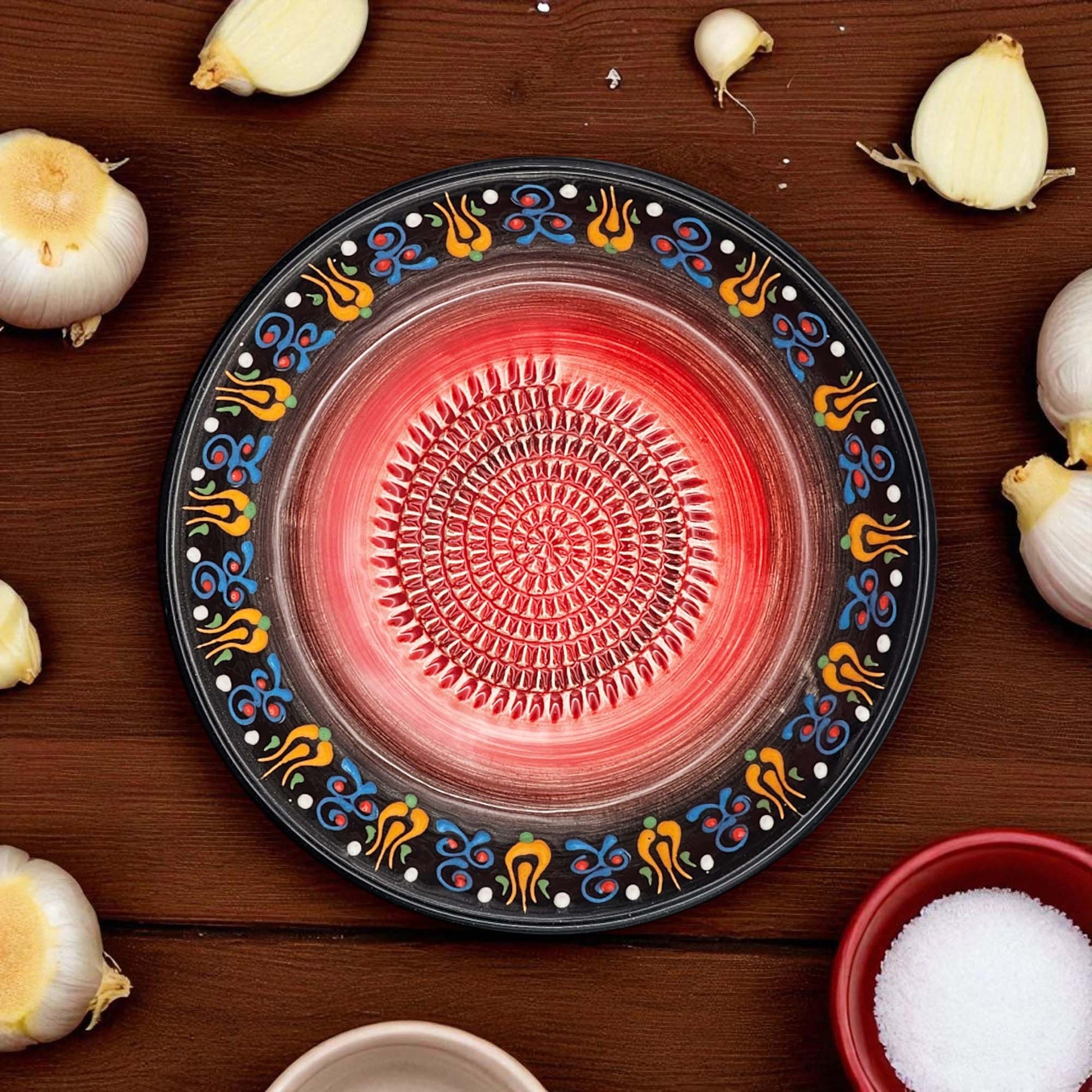 Decorative garlic grater bowl with colorful patterns on a wooden surface with garlic bulbs and a bowl of salt.