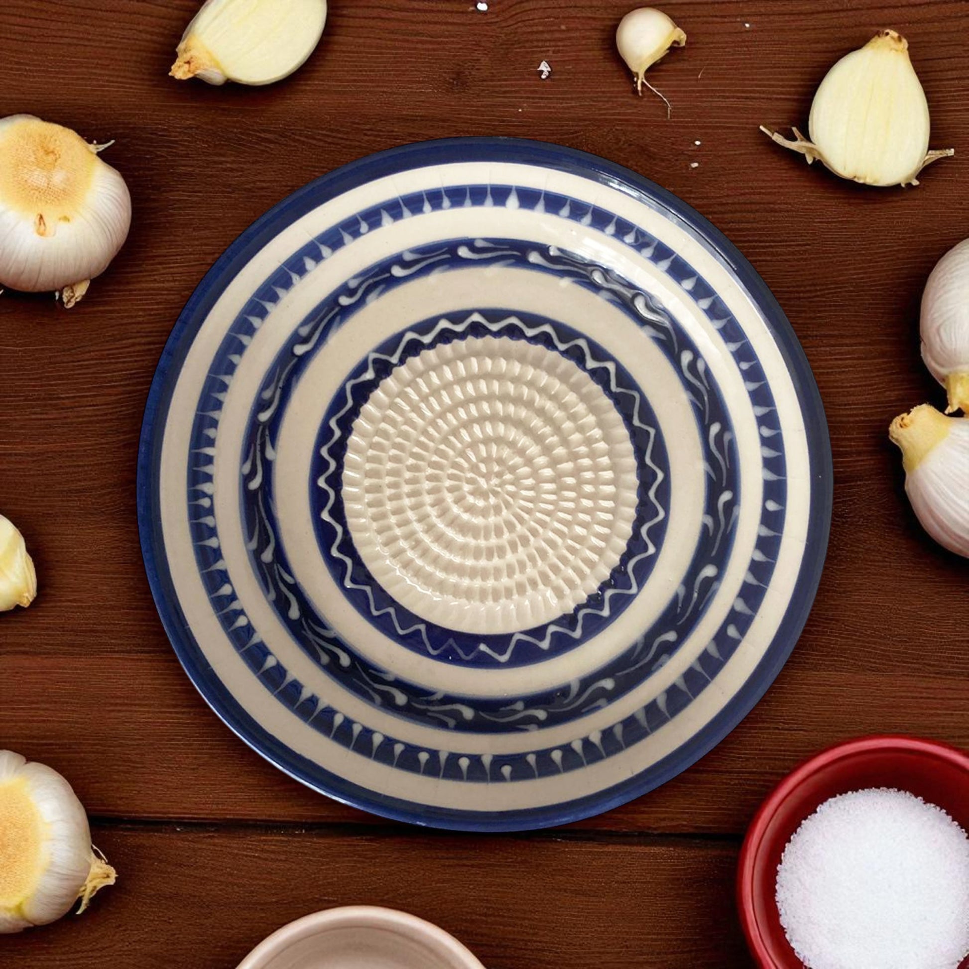 Decorative blue and white garlic grater bowl on a wooden surface with garlic bulbs and a bowl of salt.