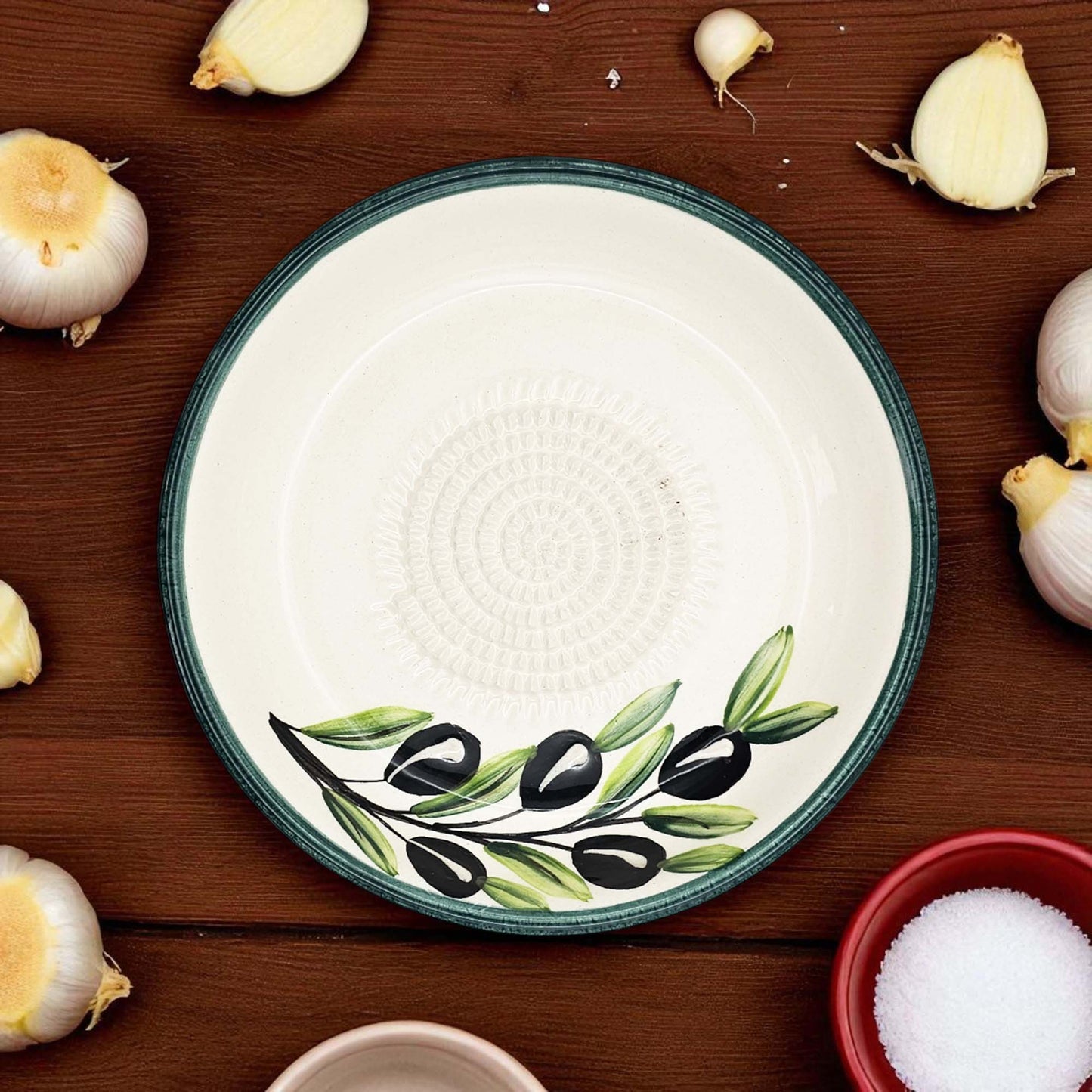 Ceramic garlic grater bowl with olive design on a wooden surface with garlic bulbs and a bowl of salt.