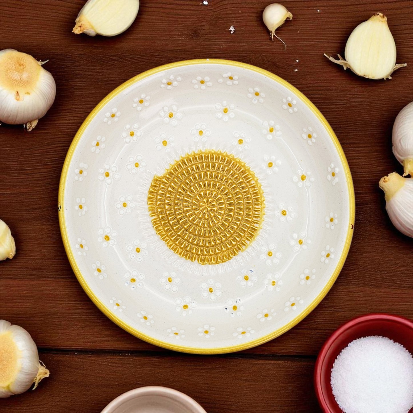 Decorative ceramic garlic grater bowl with yellow center and white rim on a wooden surface with garlic bulbs.
