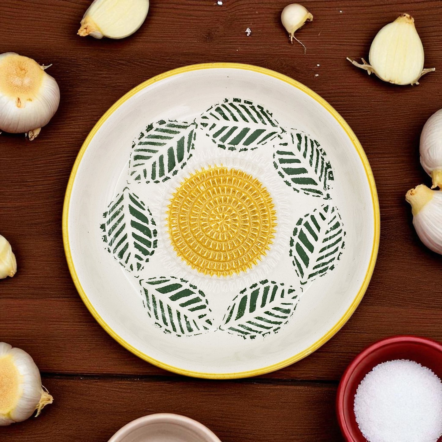 Decorative garlic grater bowl with a yellow center and green leaf patterns on a wooden surface with garlic bulbs.