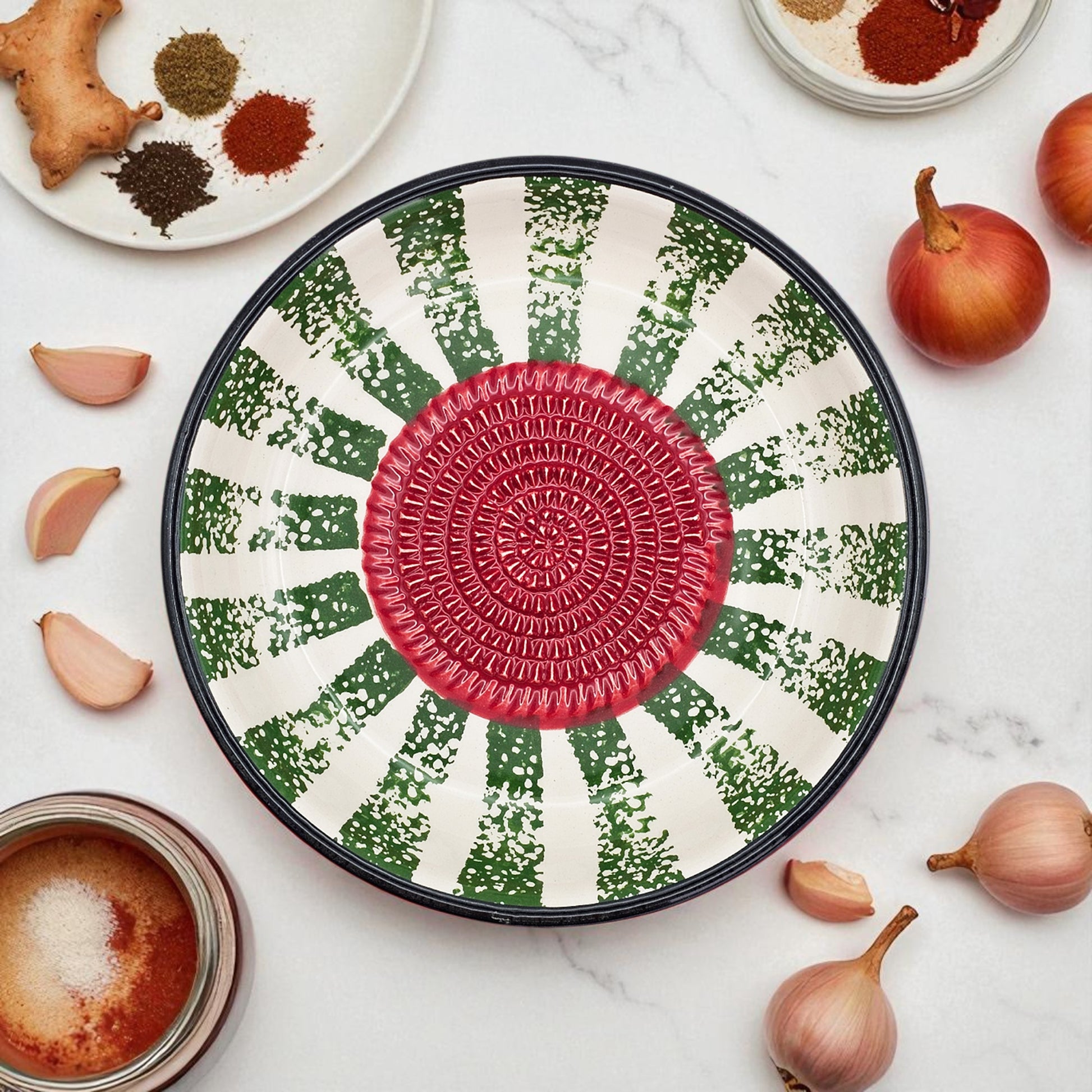 Decorative garlic grater bowl with a red center and green and white pattern on a marble surface with ingredients.