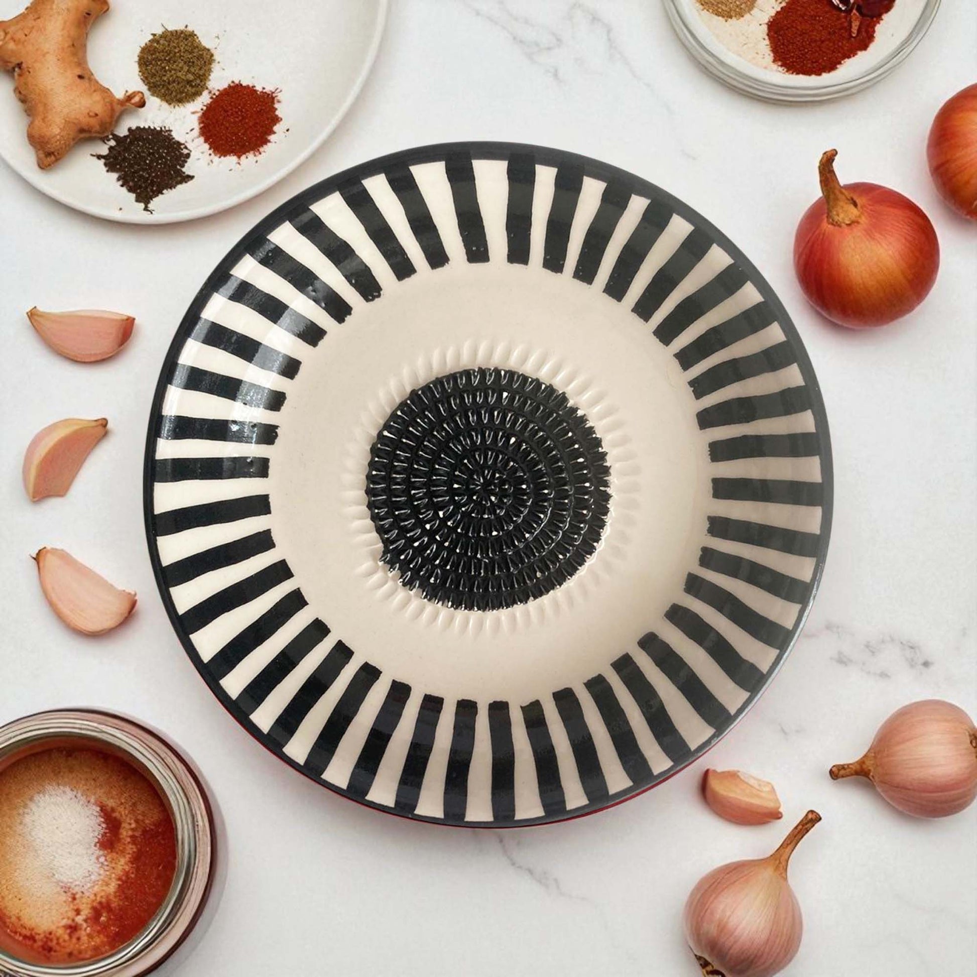 Black and white striped ceramic garlic grater bowl on a marble surface with ingredients around it.