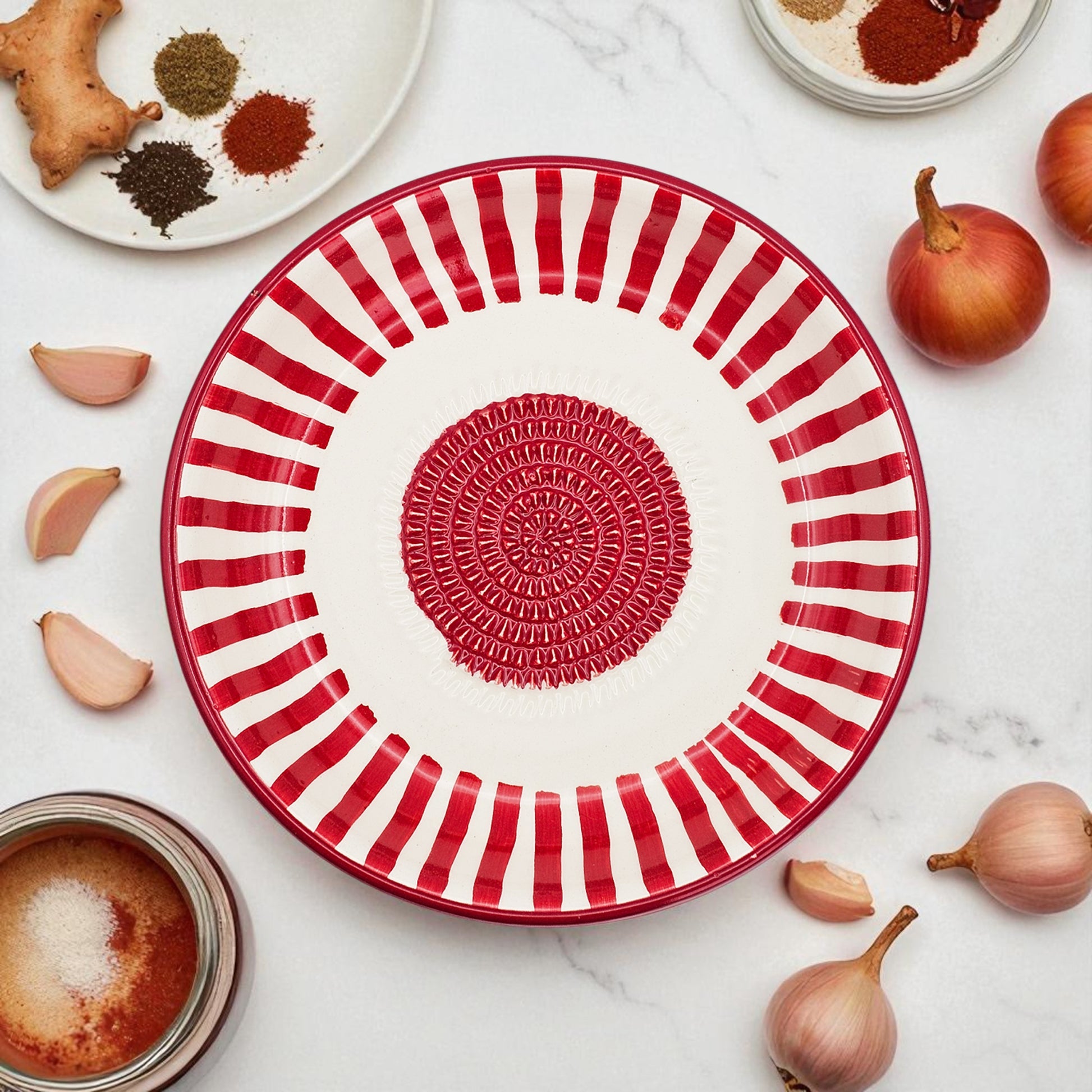 Red and white striped garlic grater bowl with decorative center on a marble surface with ingredients.