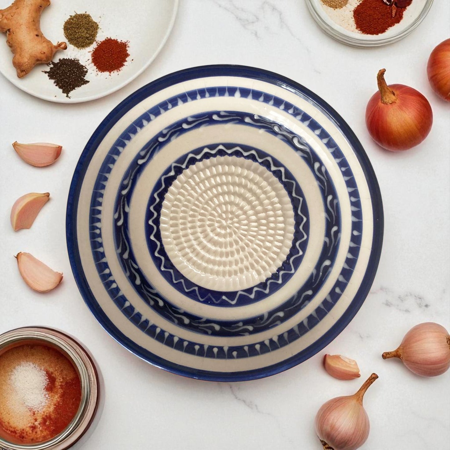 Decorative blue and white ceramic garlic grater bowl on a marble surface with spices and ingredients.