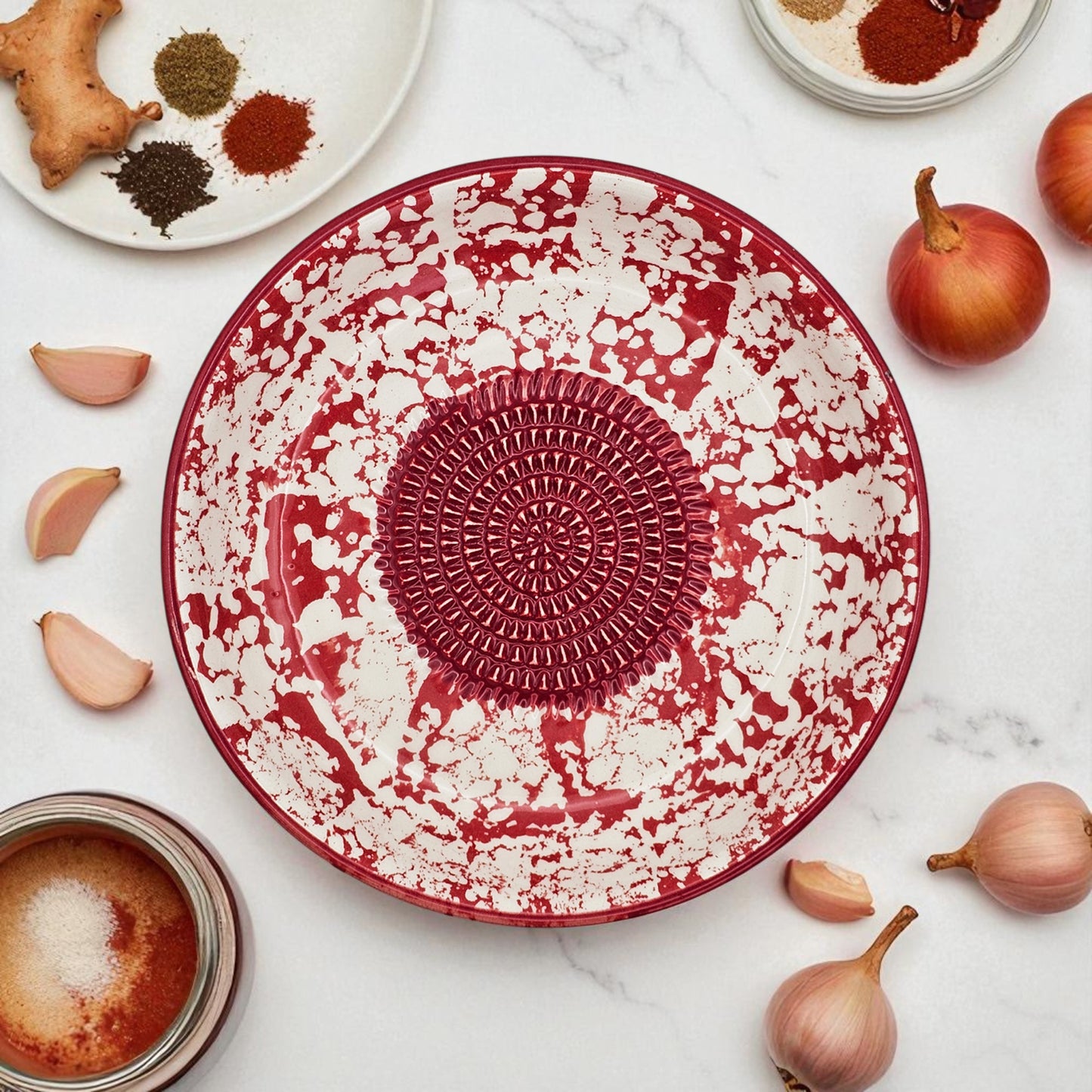 Red and white decorative garlic grater bowl on a marble surface with ingredients around it.