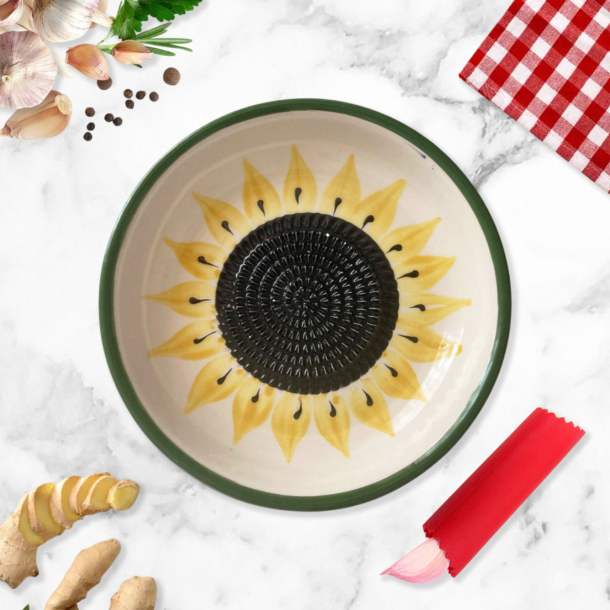 Ceramic garlic grater bowl with sunflower design on a marble surface with ginger and a red checkered cloth.