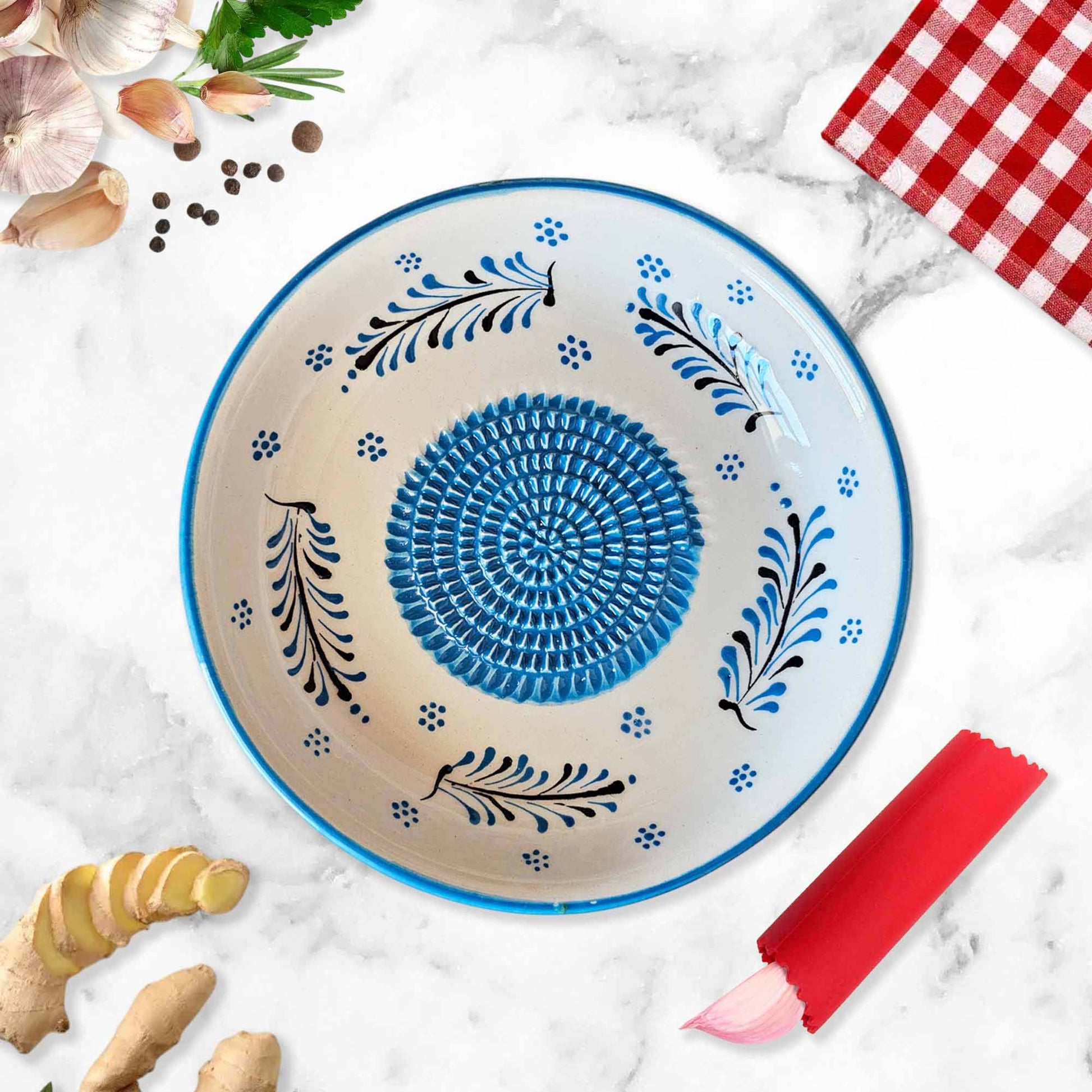 Decorative ceramic garlic grater bowl with blue center and floral patterns on a marble surface with ginger and a red checkered cloth.