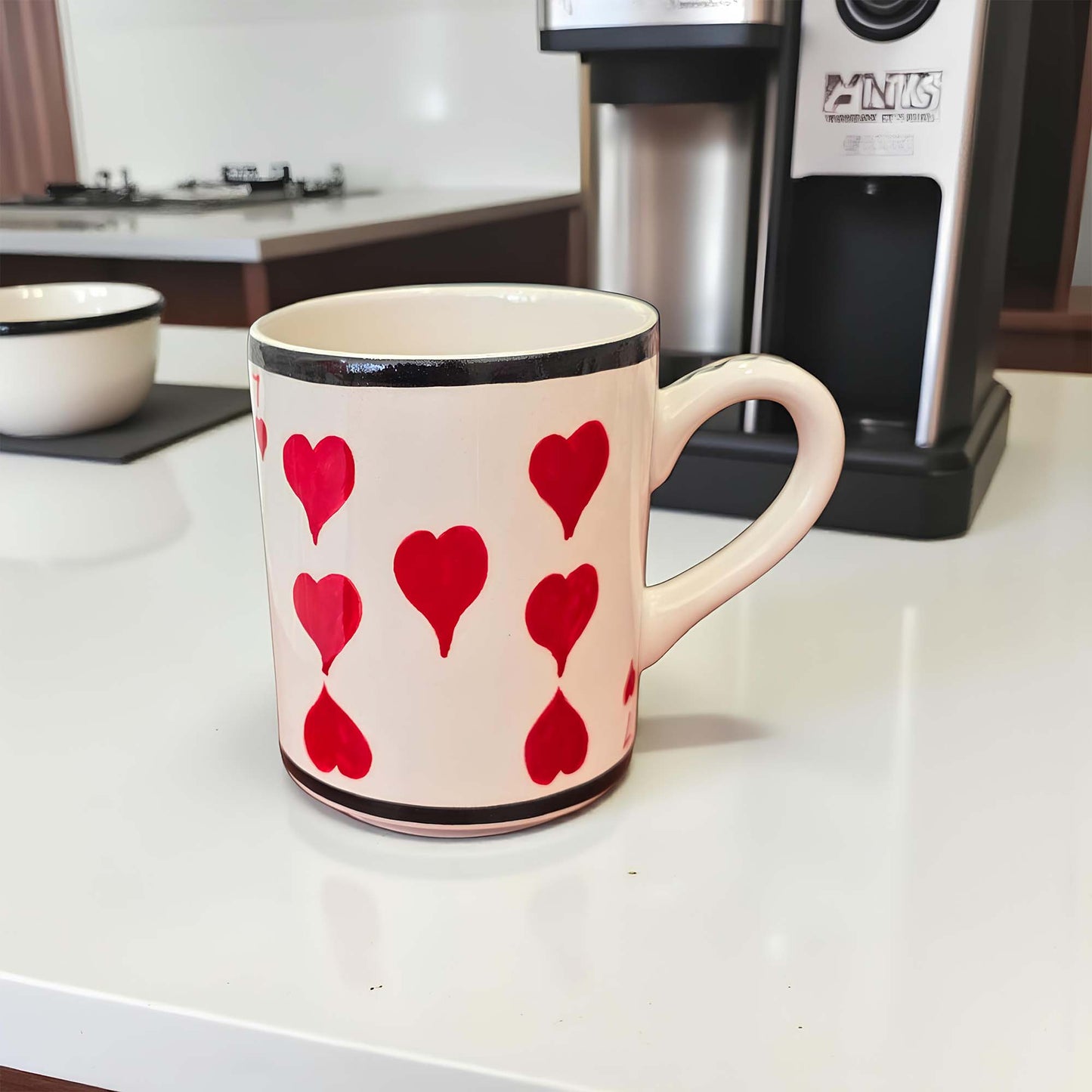 Mug with red heart patterns on a kitchen counter with a coffee maker in the background.