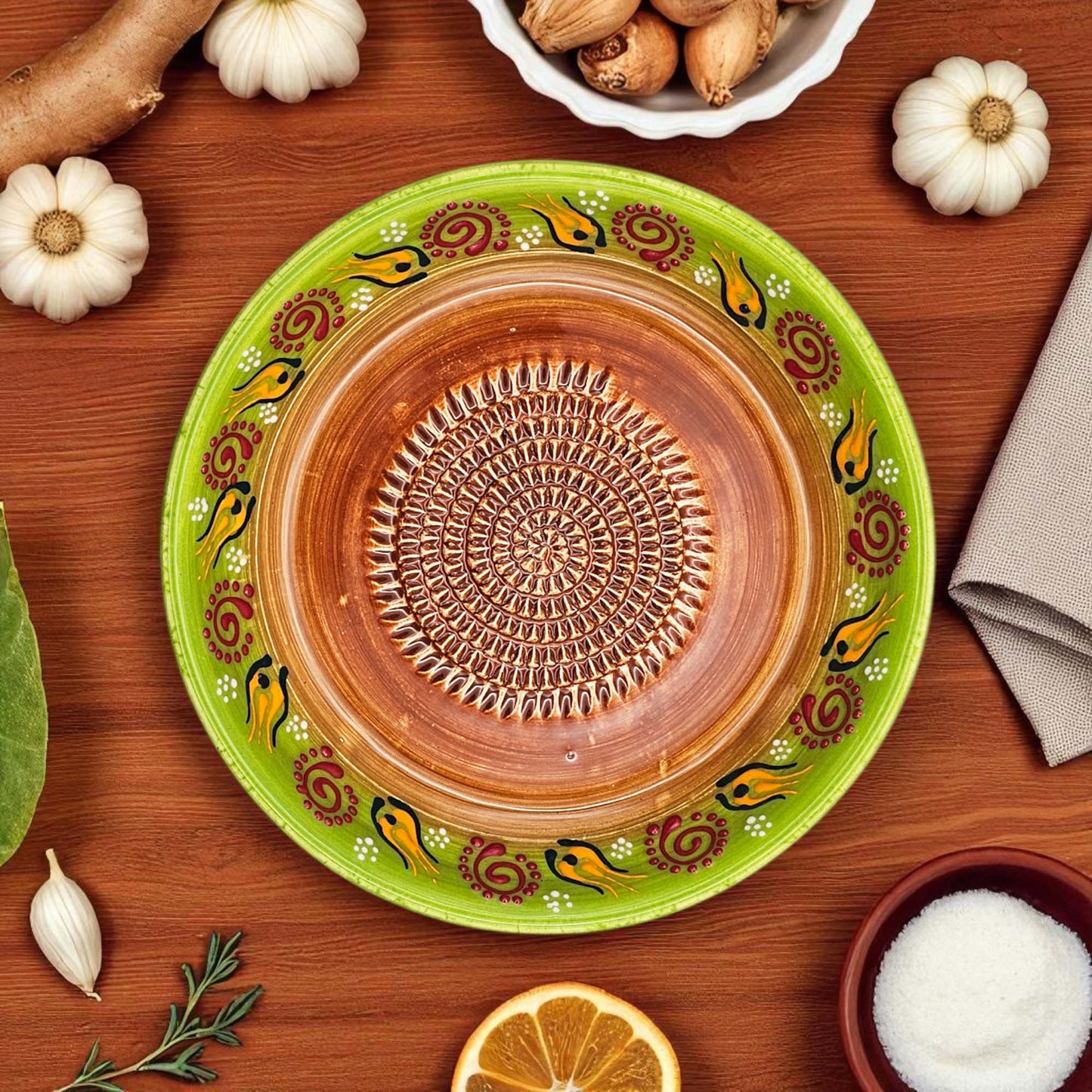 Decorative ceramic garlic grater bowl with green rim and brown center on a wooden surface with garlic and oranges.