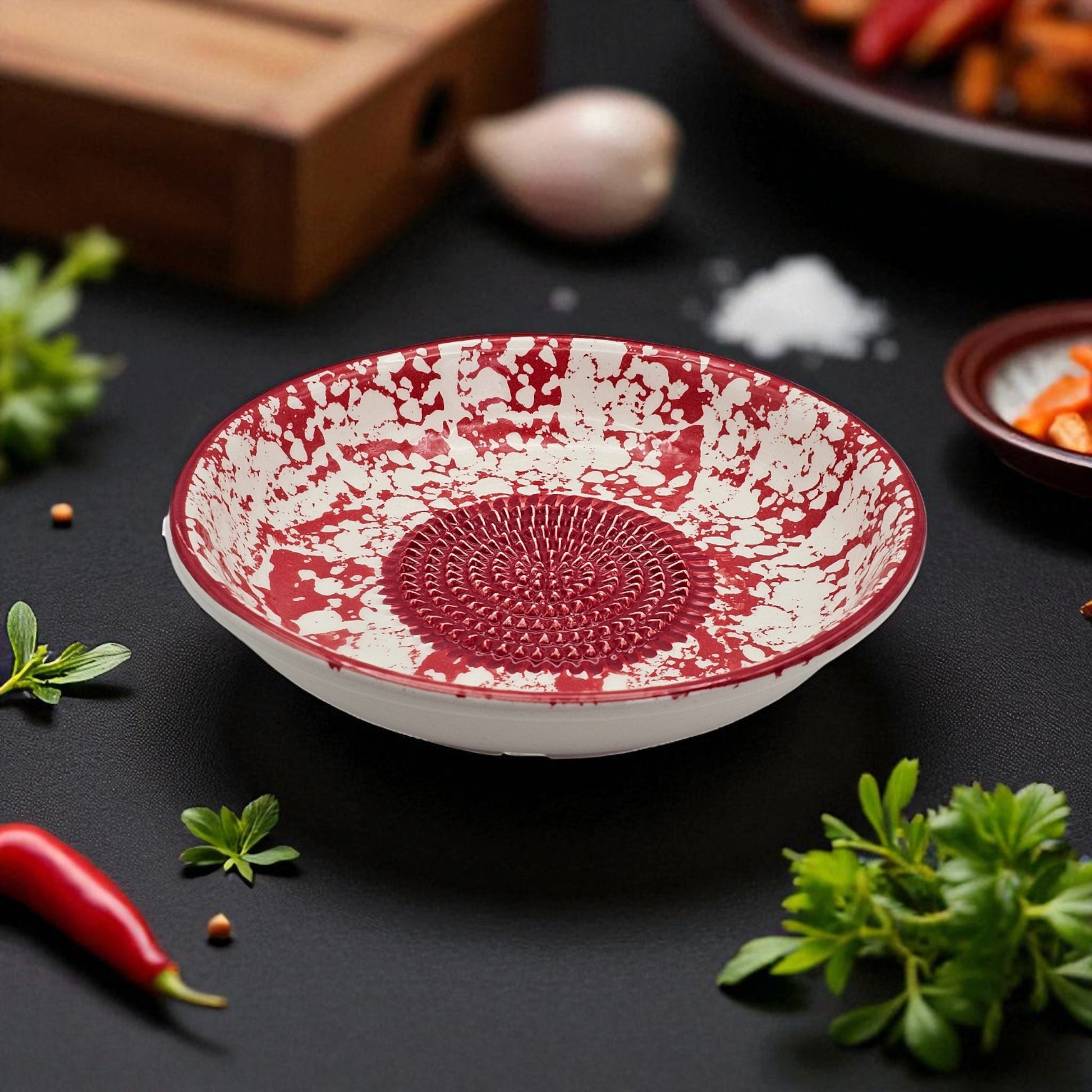 Red and white textured garlic grater bowl on a dark surface with herbs and spices.