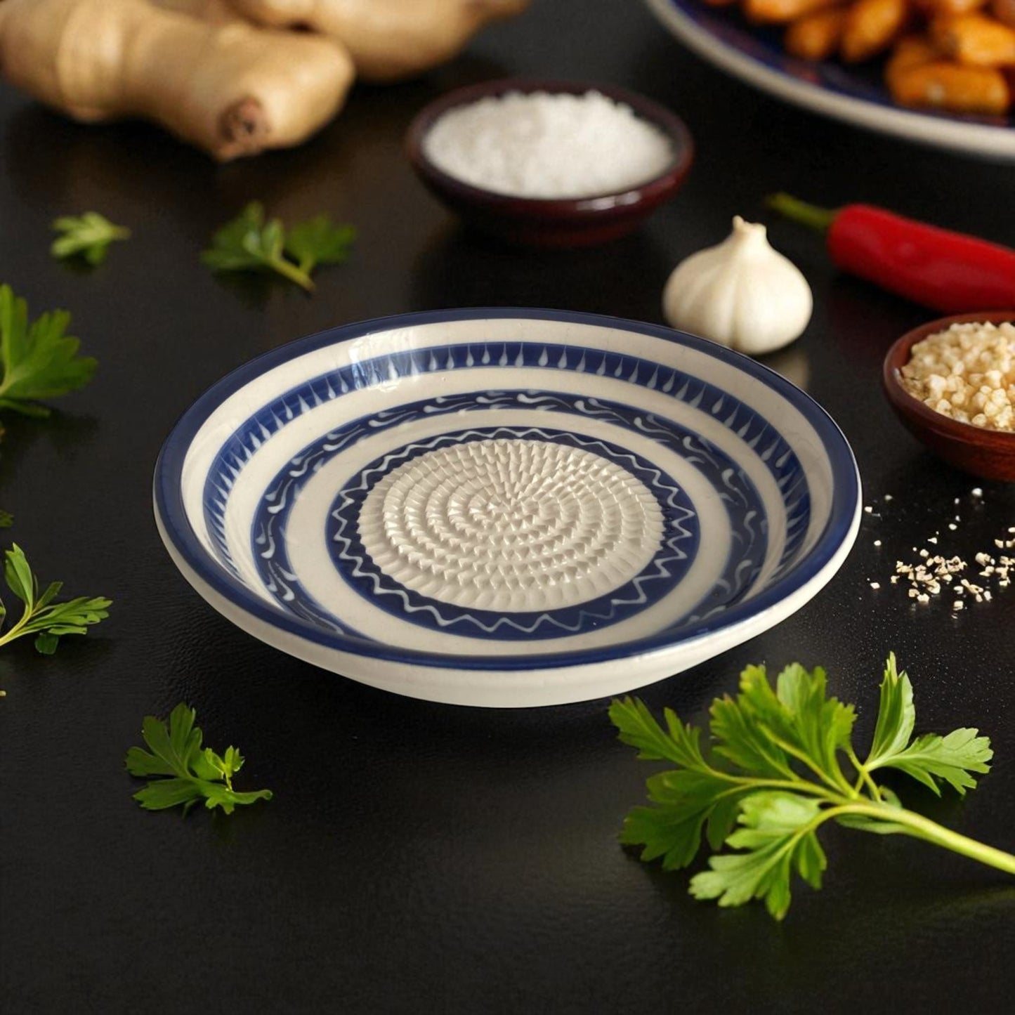 Blue and white decorative garlic grater bowl on a dark surface with herbs and spices.