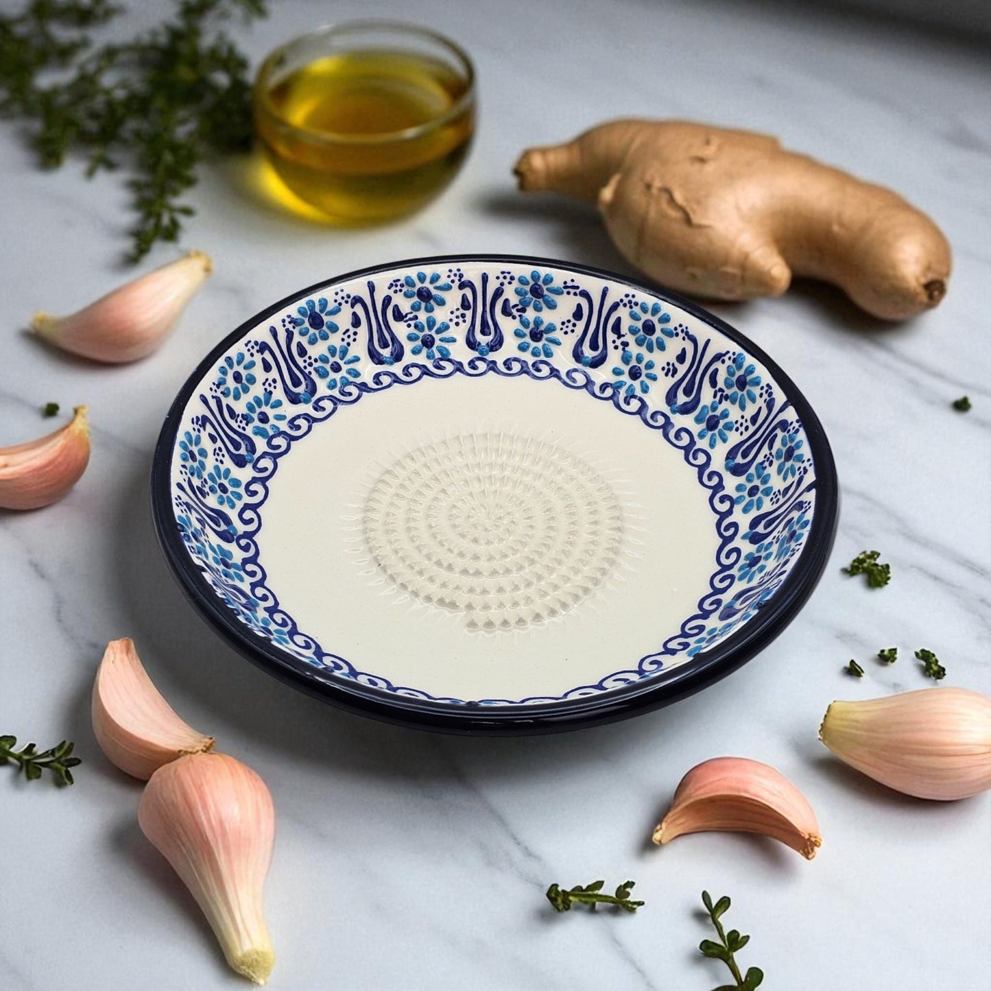 Decorative ceramic garlic grater plate with blue floral patterns on a marble surface with garlic and olive oil.