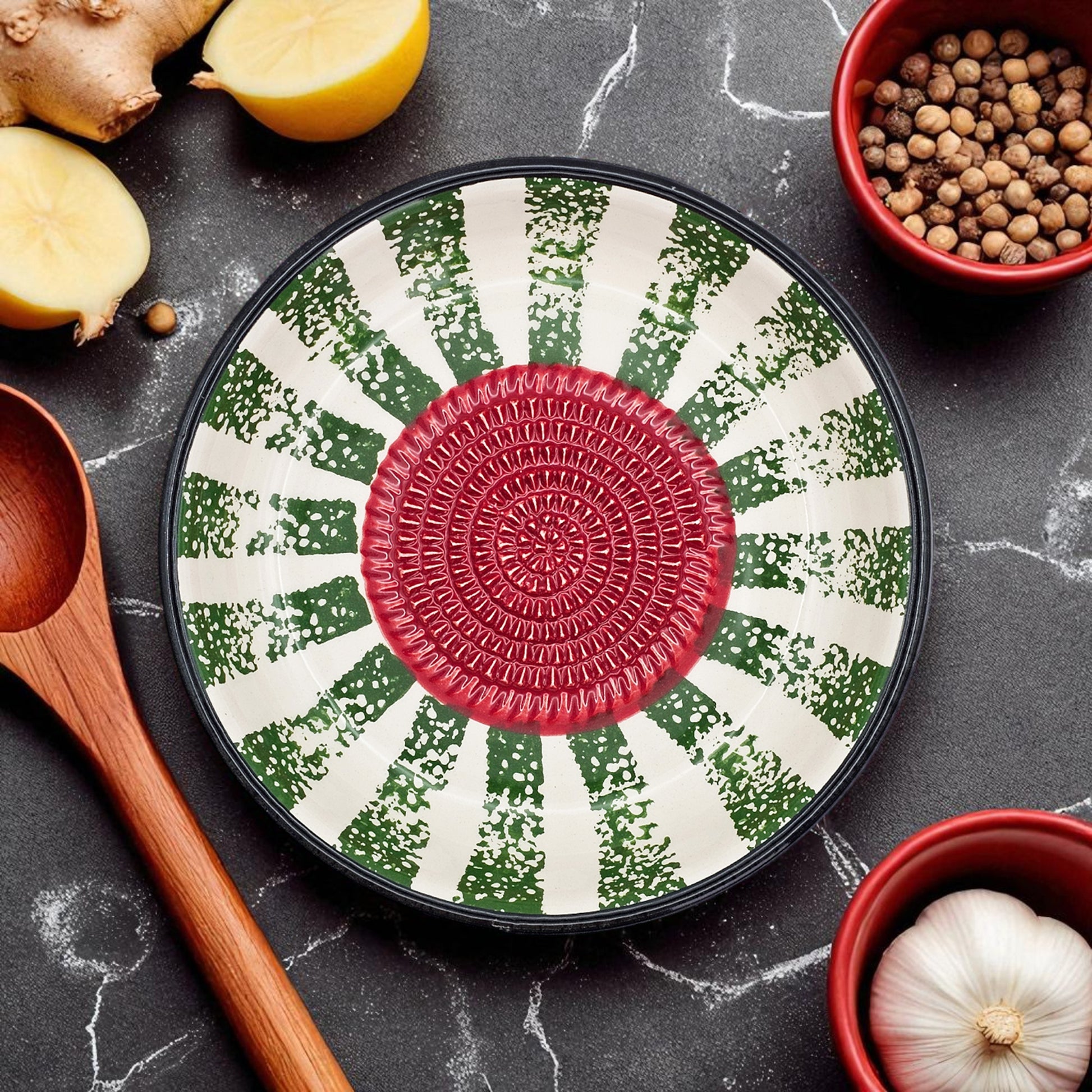 Decorative garlic grater bowl with a red center and green and white pattern on a dark surface with kitchen items.