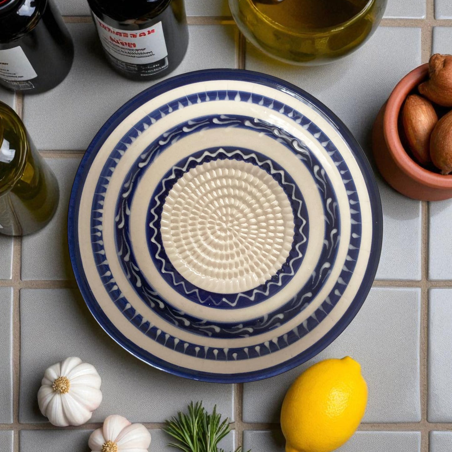 Blue and white ceramic garlic grater bowl on a tiled surface with ingredients like lemons and garlic.