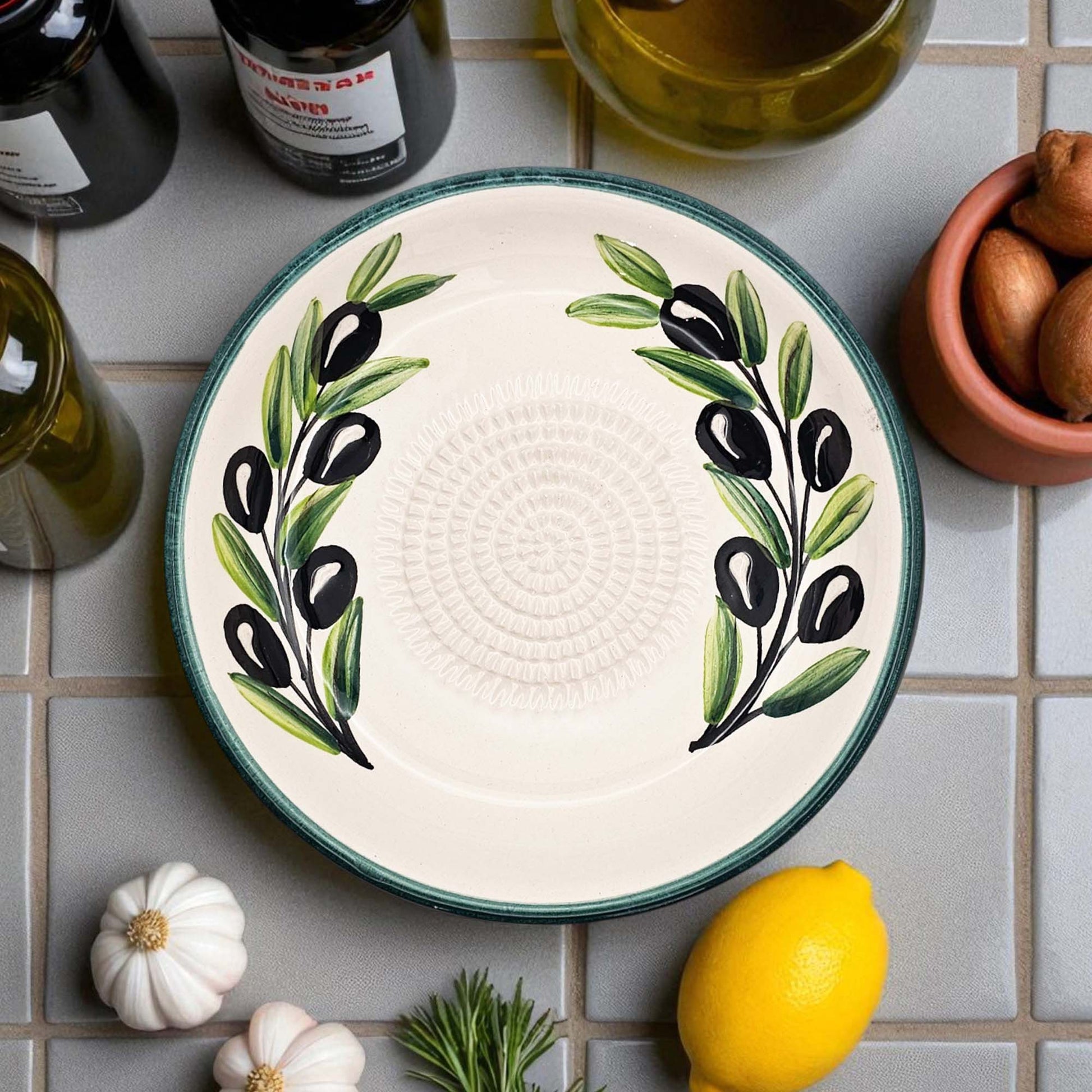 Decorative garlic grater bowl with olive design on a tiled surface with ingredients.