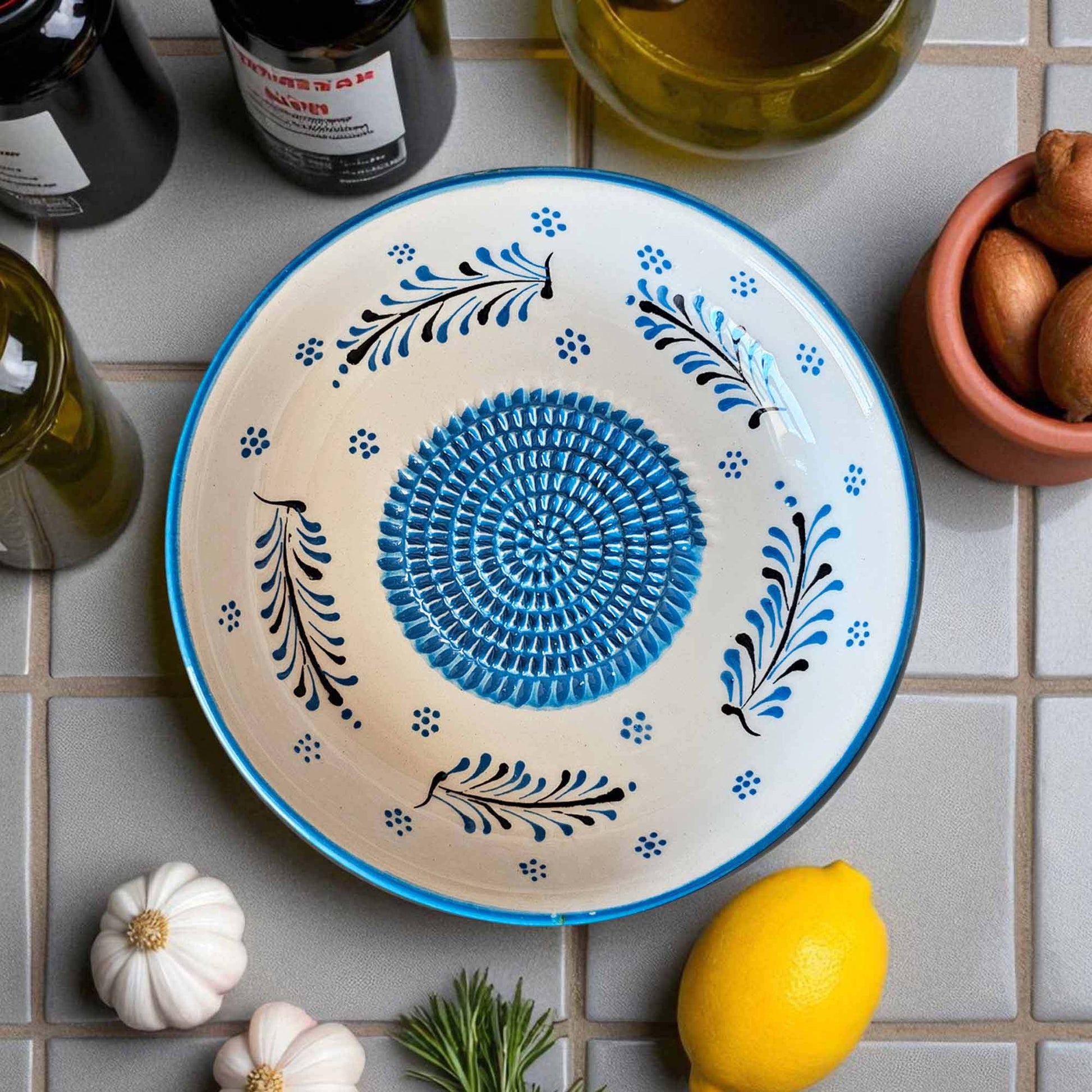 Decorative garlic grater bowl with blue and black patterns on a tiled surface with lemons and bottles.