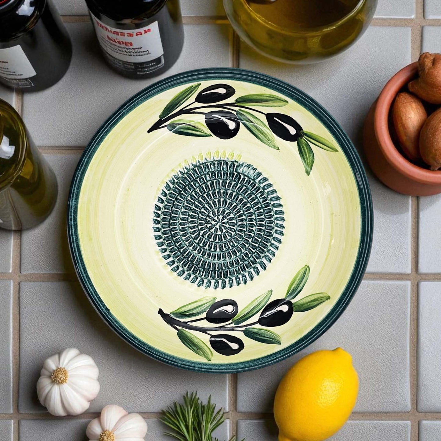 Decorative ceramic garlic grater bowl with olive design on a tiled surface with ingredients.
