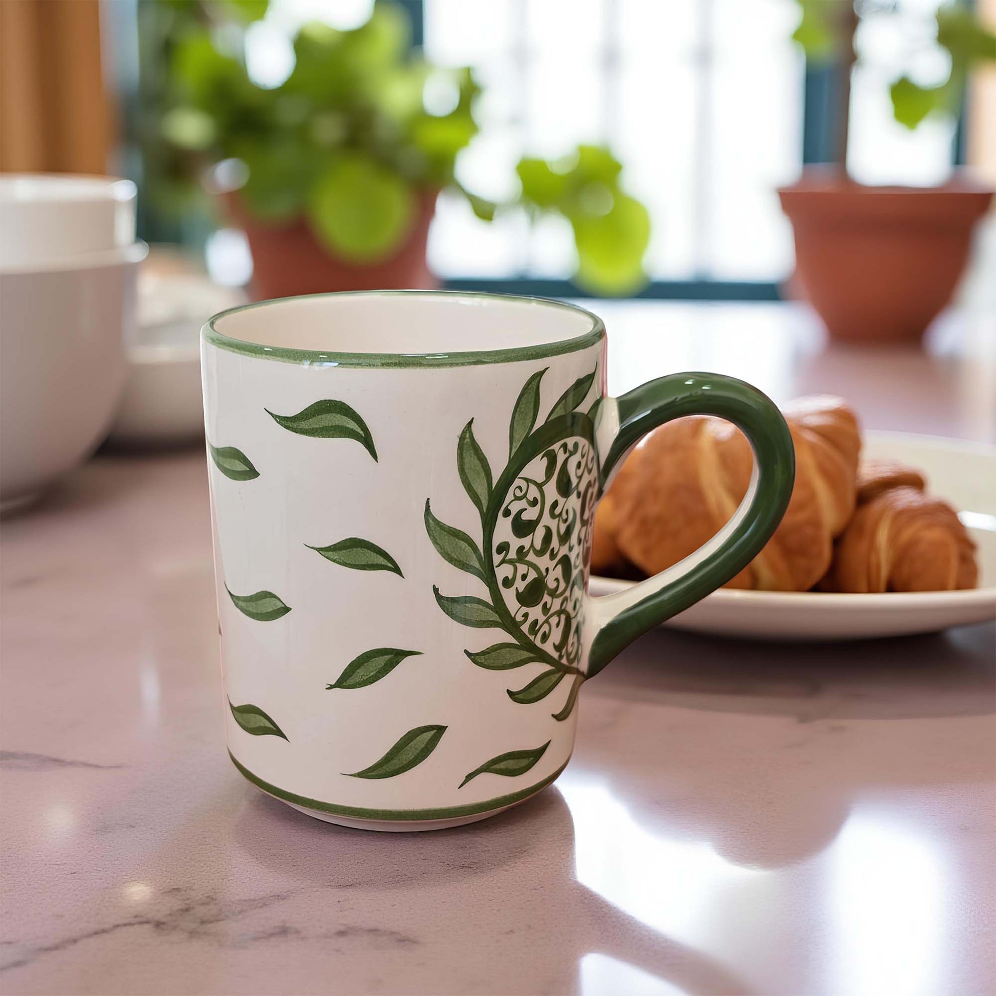 White mug with green leaf pattern on a marble surface with pastries and plants in the background