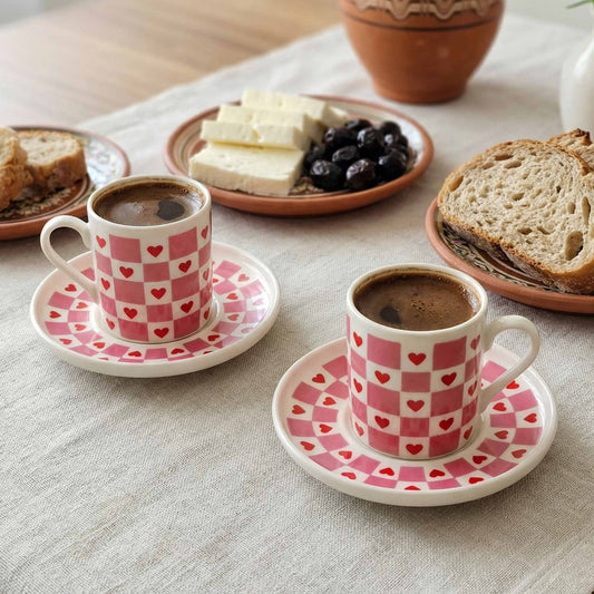 Two pink checkered coffee cups with saucers on a table with bread and cheese.