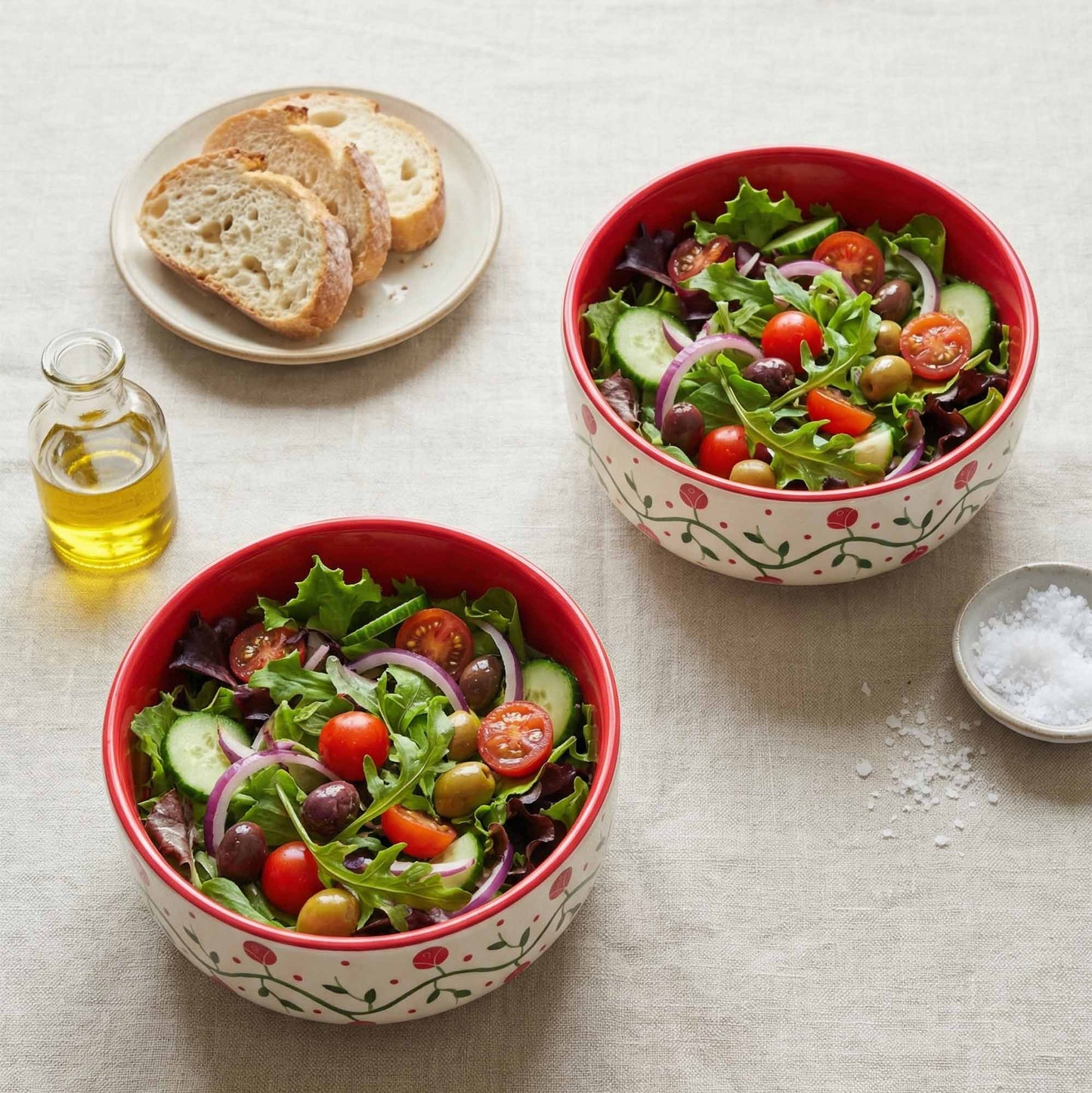 Two red bowls of salad with bread and olive oil on a light surface