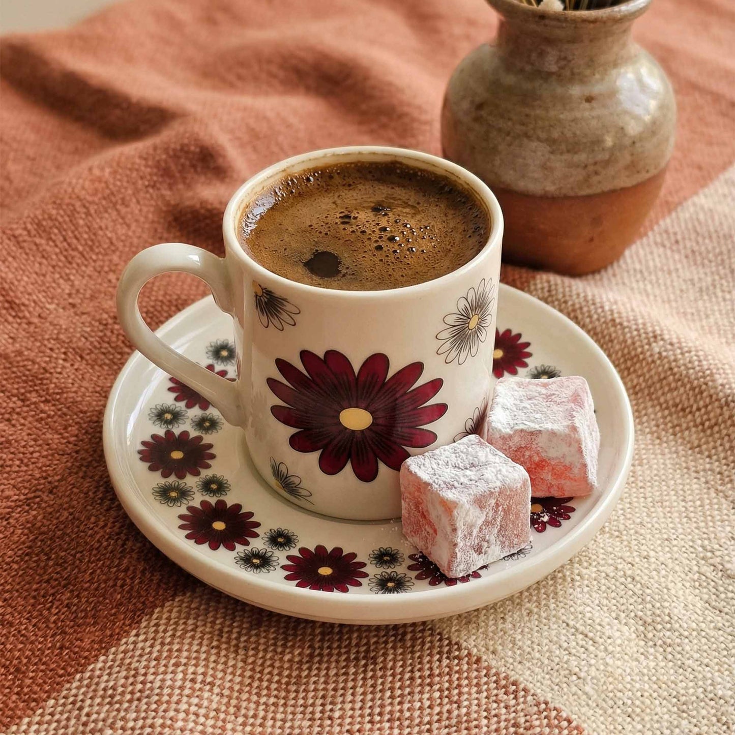 Cup of coffee with a floral design on a saucer, accompanied by Turkish delight, on a textured fabric background.