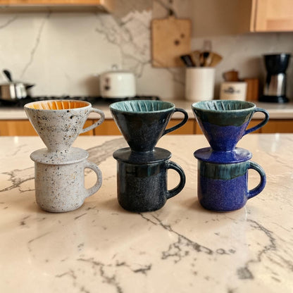 Four ceramic coffee filters and mugs stacked on a marble countertop with a kitchen background.