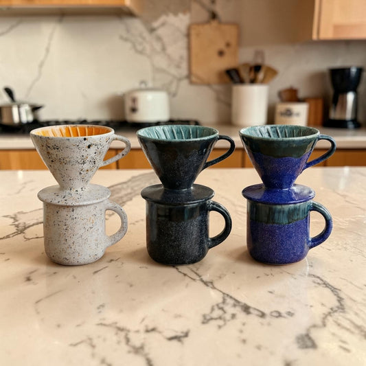 Four ceramic coffee filters and mugs stacked on a marble countertop with a kitchen background.