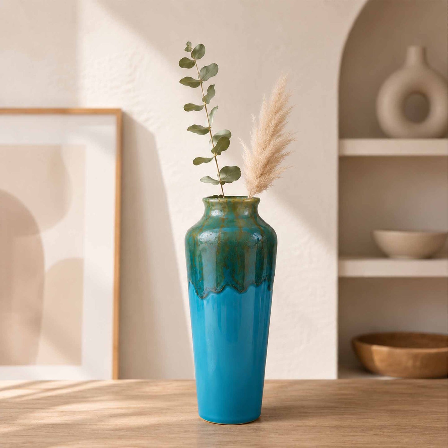 Ceramic vase with plant and dried elements on a wooden surface, with a neutral background.