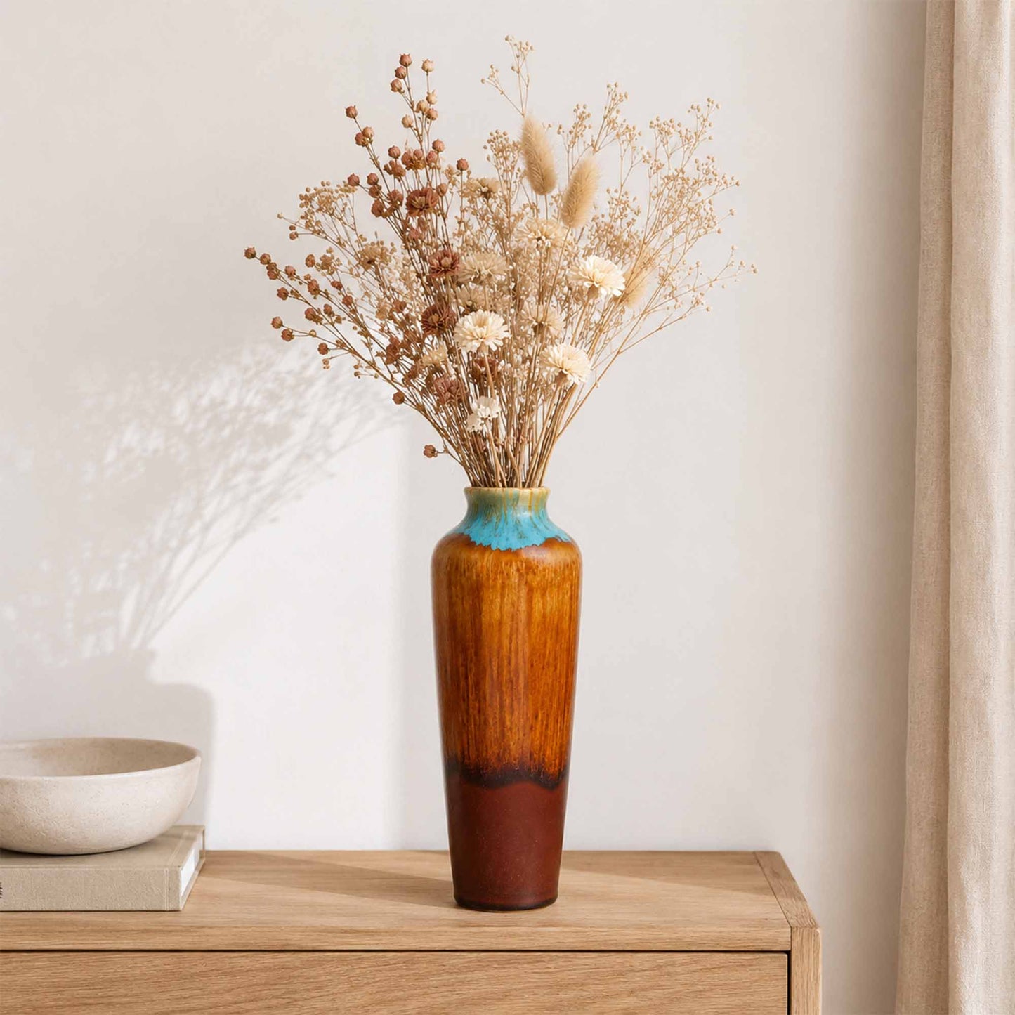 Tall ceramic vase with dried flowers on a wooden surface against a neutral wall.