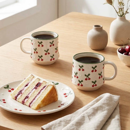 Two mugs with cherry design on a table with a slice of cake and a vase.