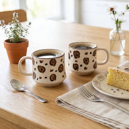 Two mugs with coffee seeds pattern on a wooden table with a slice of cake and utensils.