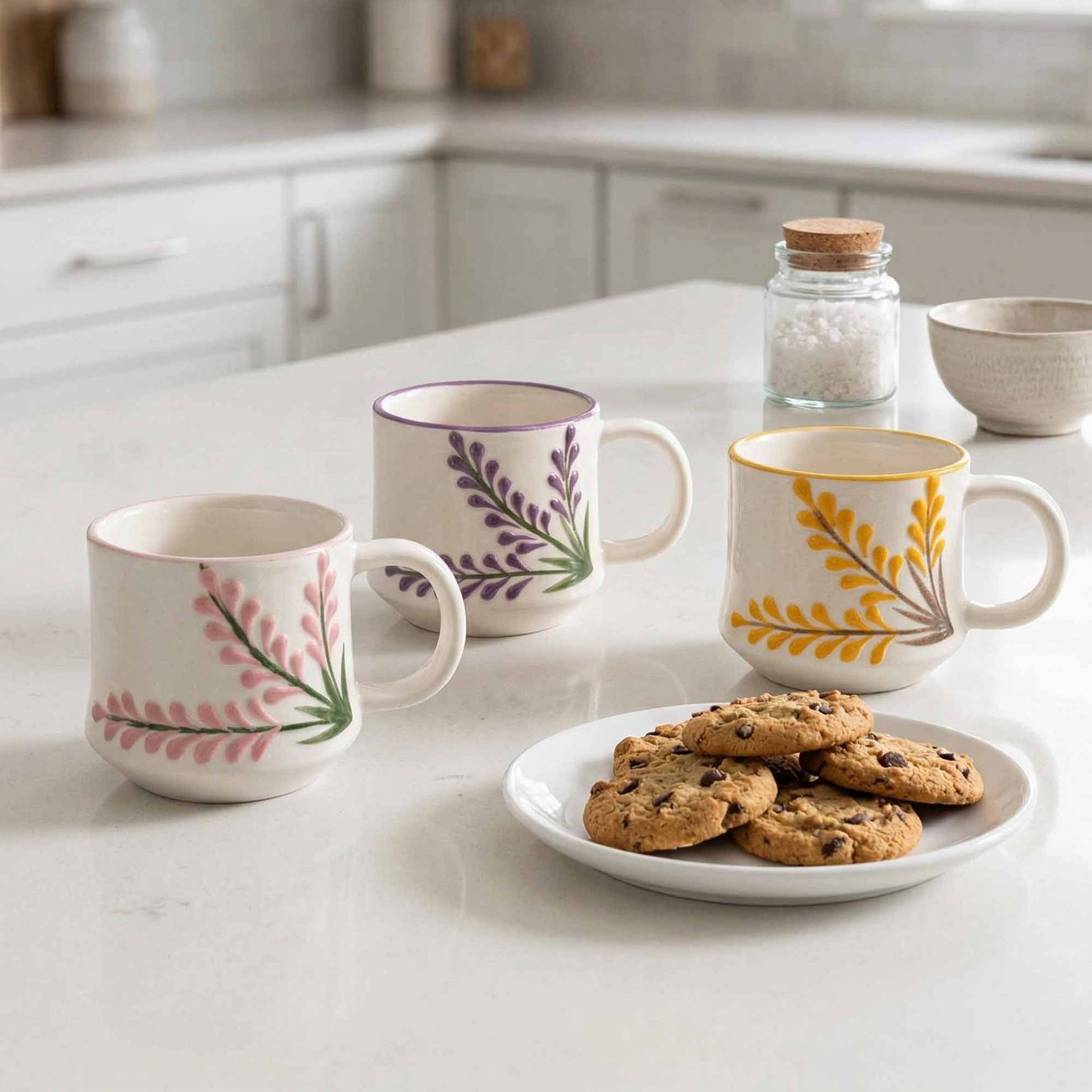 Three mugs with floral designs on a kitchen counter with a plate of cookies.