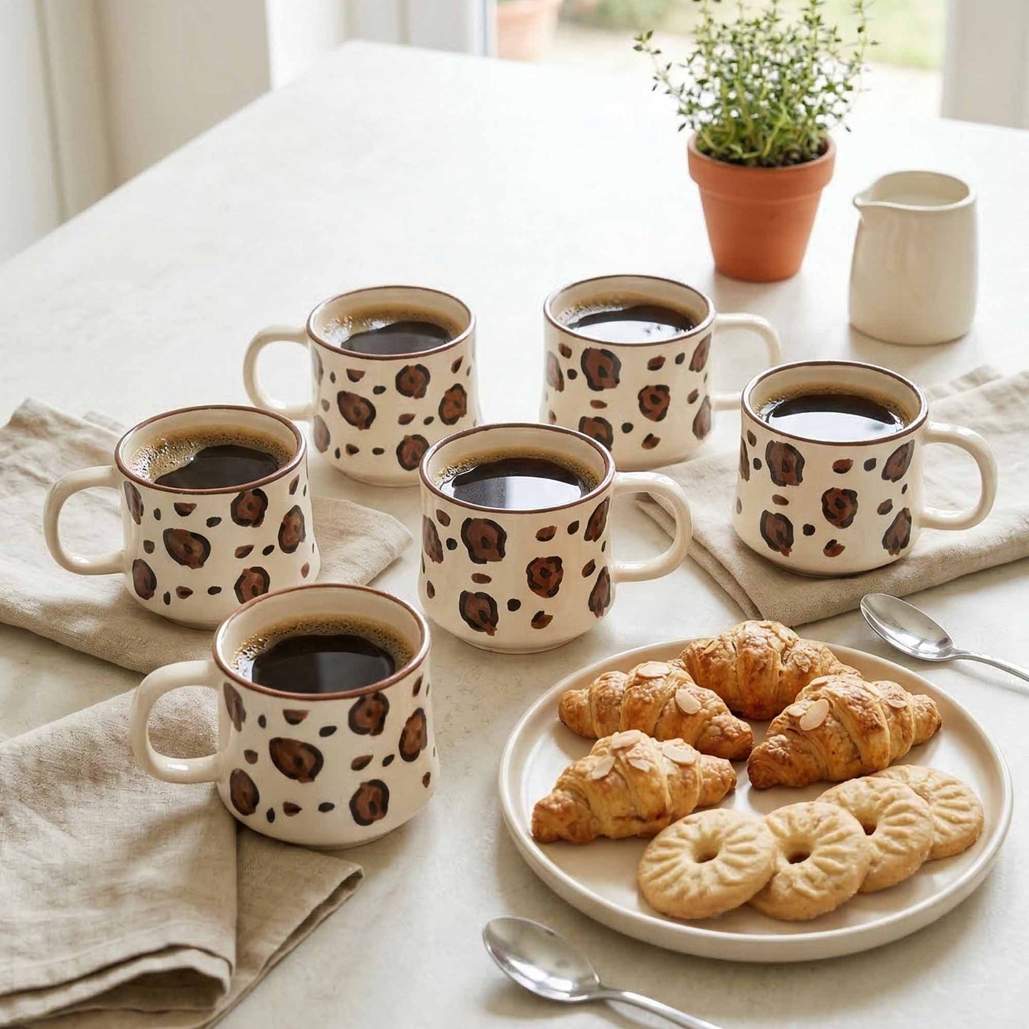 Set of mugs with coffee seeds print pattern on a table with pastries and a plant.
