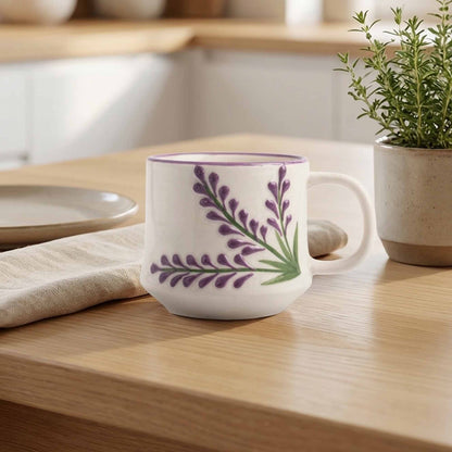 White mug with purple floral design on a wooden table with a plant and plates in the background