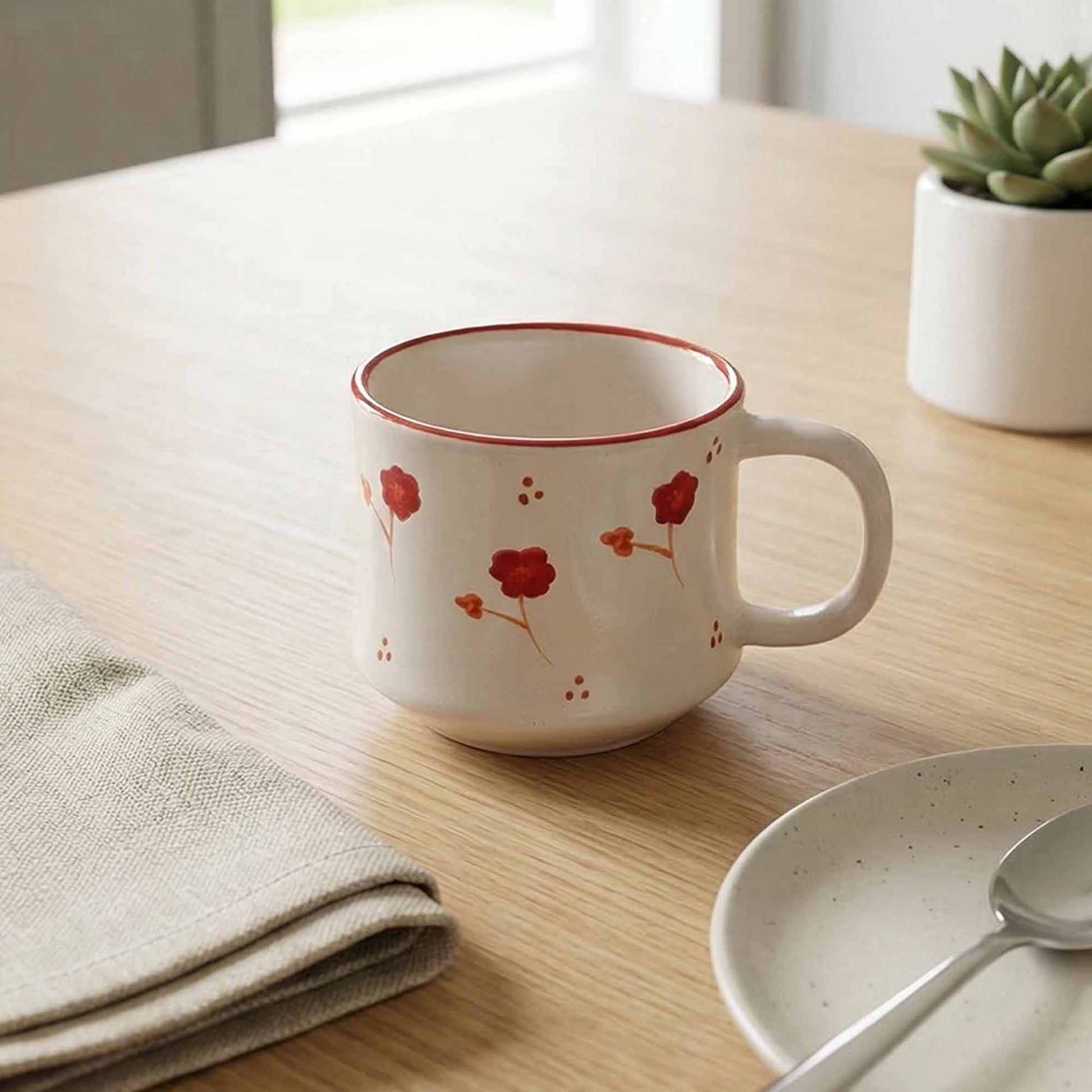 White mug with red floral design on a wooden table with a plant and folded cloth in the background