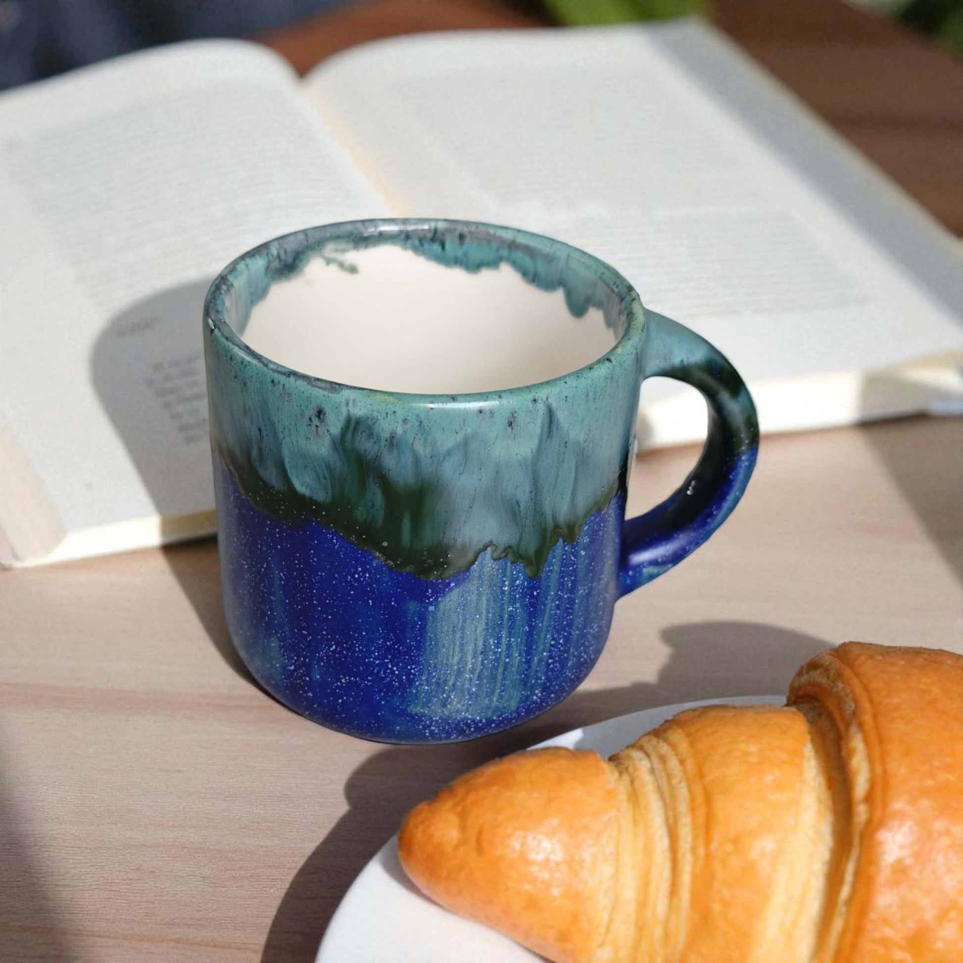 Blue ceramic mug with a white interior, placed on a wooden surface next to a croissant and an open book.