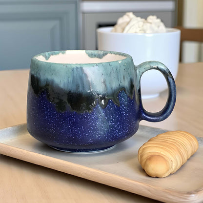 Blue ceramic mug with a handle on a wooden tray, with a bowl of whipped cream and a croissant in the background.