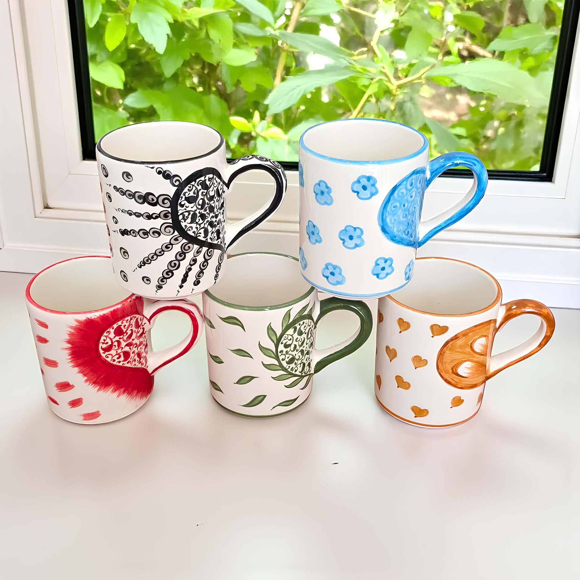 Five colorful mugs with various patterns on a windowsill with greenery outside.