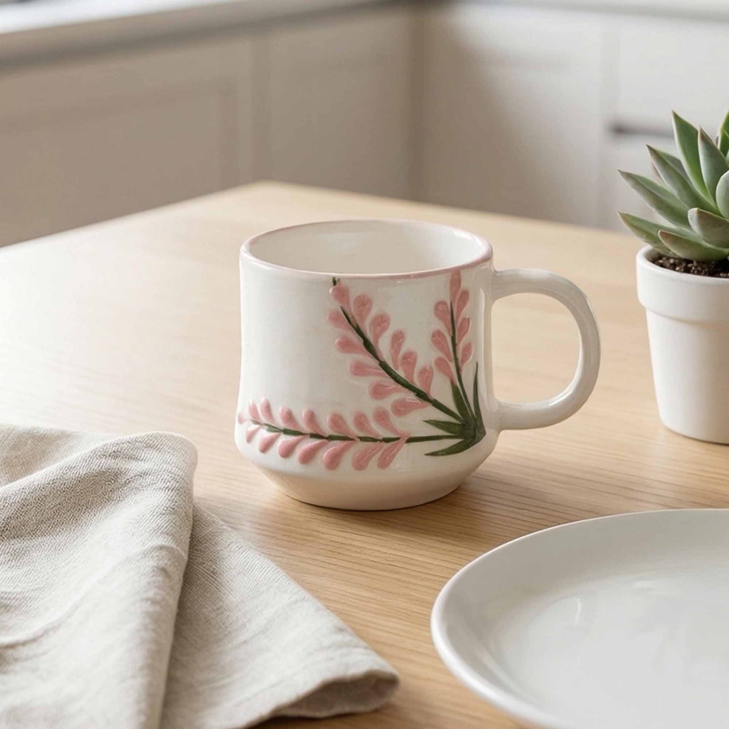 White mug with pink floral design on a wooden surface with a plant and towel.