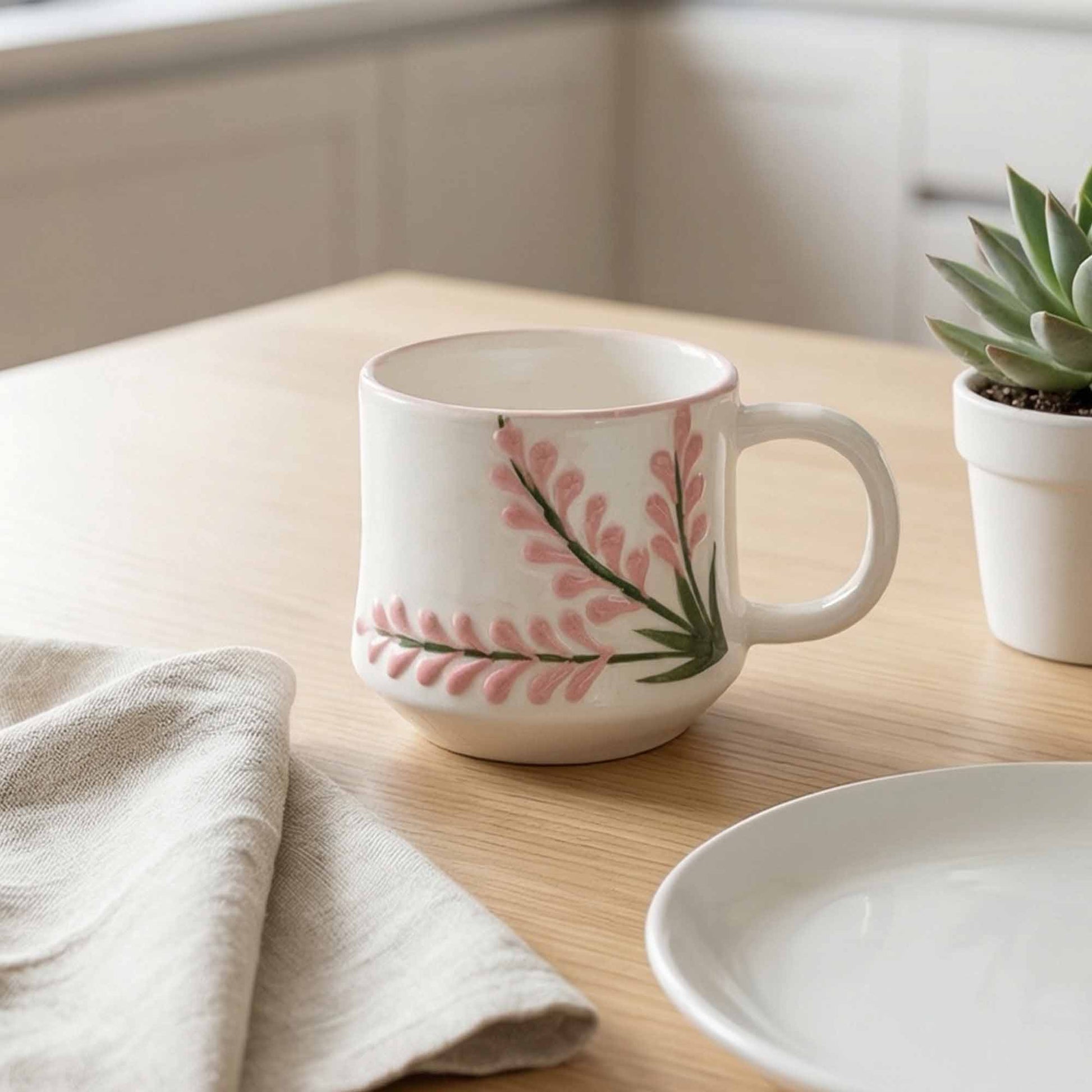 White mug with pink floral design on a wooden surface with a plant and towel.