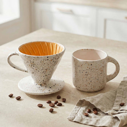 Speckled ceramic coffee filter and mug on a light surface with scattered coffee beans.