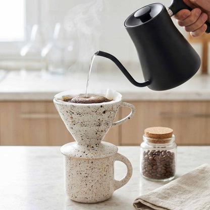 Person pouring coffee from a black kettle into a speckled ceramic coffee filter.